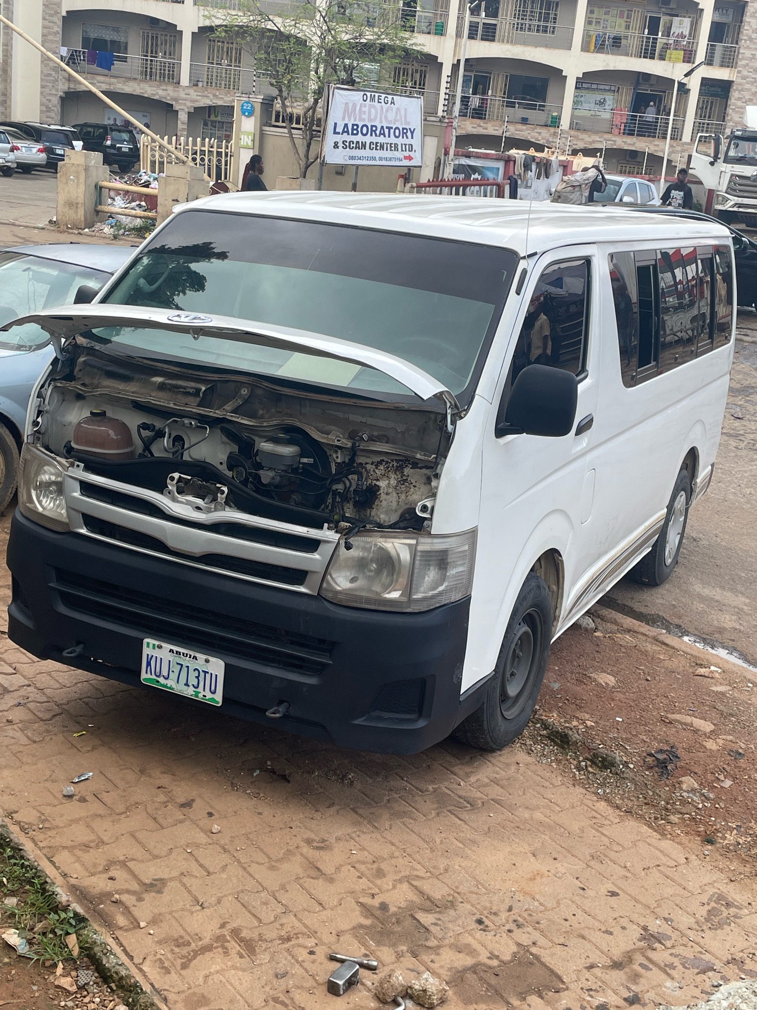 The image shows a white Toyota Hiace van with its hood open, revealing the engine compartment. The van is parked on a paved street in Abuja, Nigeria. A blue and green license plate is visible on the front of the van, reading "KUJ-713TU". In the background, a multi-story building with balconies and shops can be seen. Other vehicles are parked in front of the building, and there are some people standing around. The scene appears to be a street in a commercial or residential area.