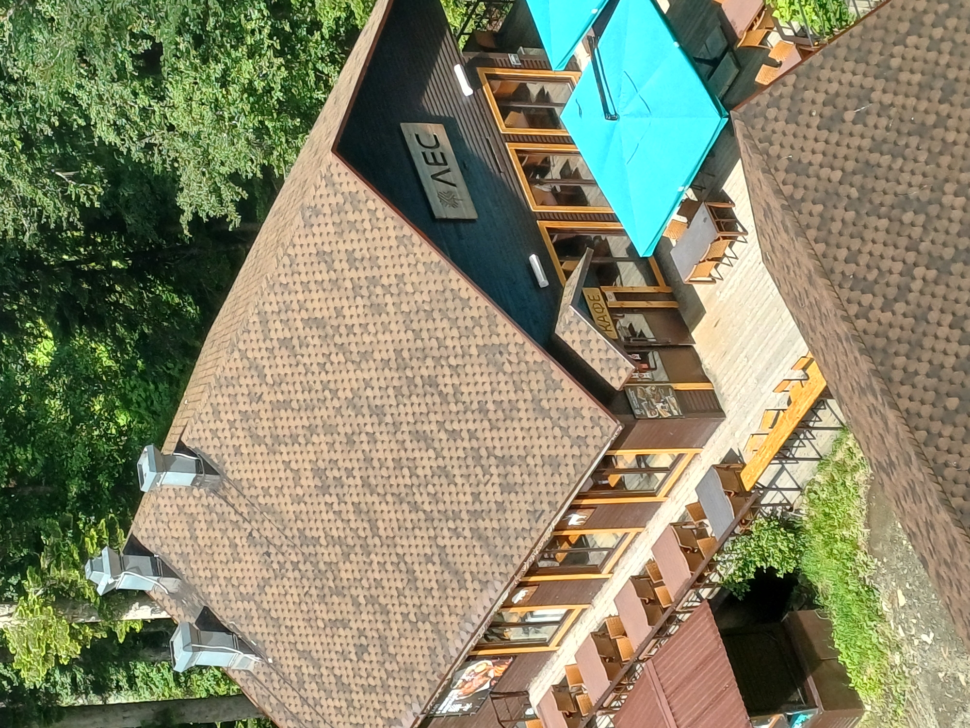 The image captures an aerial view of a restaurant or cafe complex in Estosadok, Russia, on a sunny day. The architecture features wooden structures with brown shingled roofs, and a prominent dark blue facade on the main building. Several outdoor seating areas are visible, furnished with tables and chairs. Two large, bright turquoise umbrellas provide shade over a patio area. One building has a sign with the Cyrillic letters "АЕС" and a stylized tree logo. Another entrance displays the word "КАФЕ" in Cyrillic. Surrounding the buildings are lush green trees, indicative of a natural or park-like setting. The image appears to be taken from a high vantage point, possibly a drone or a higher building.