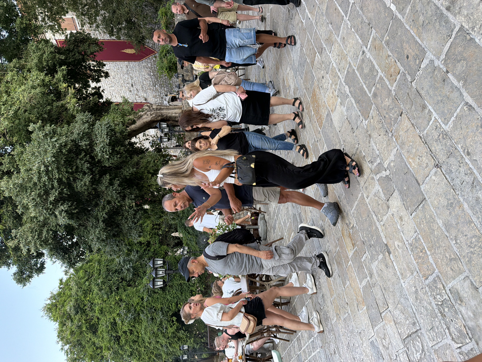 The image captures a scene in the city of Budva, Montenegro, specifically within the old town, likely on a paved courtyard area. A group of people are walking across a stone-paved surface. To the left, a woman in a white top and black skirt is walking and looking to her left. A man in a grey t-shirt and cargo pants, wearing a backpack, is walking past her. To the right of the man, a woman with blonde hair in a white tank top and black pants is walking, holding a black handbag. Further to the right, a man in a black t-shirt and denim shorts is walking and gesturing with his right hand. In the background, there are trees, stone buildings with red banners, and lampposts. Some people are seated at tables in what appears to be an outdoor seating area. The lighting suggests it is daytime, possibly late afternoon or early evening.