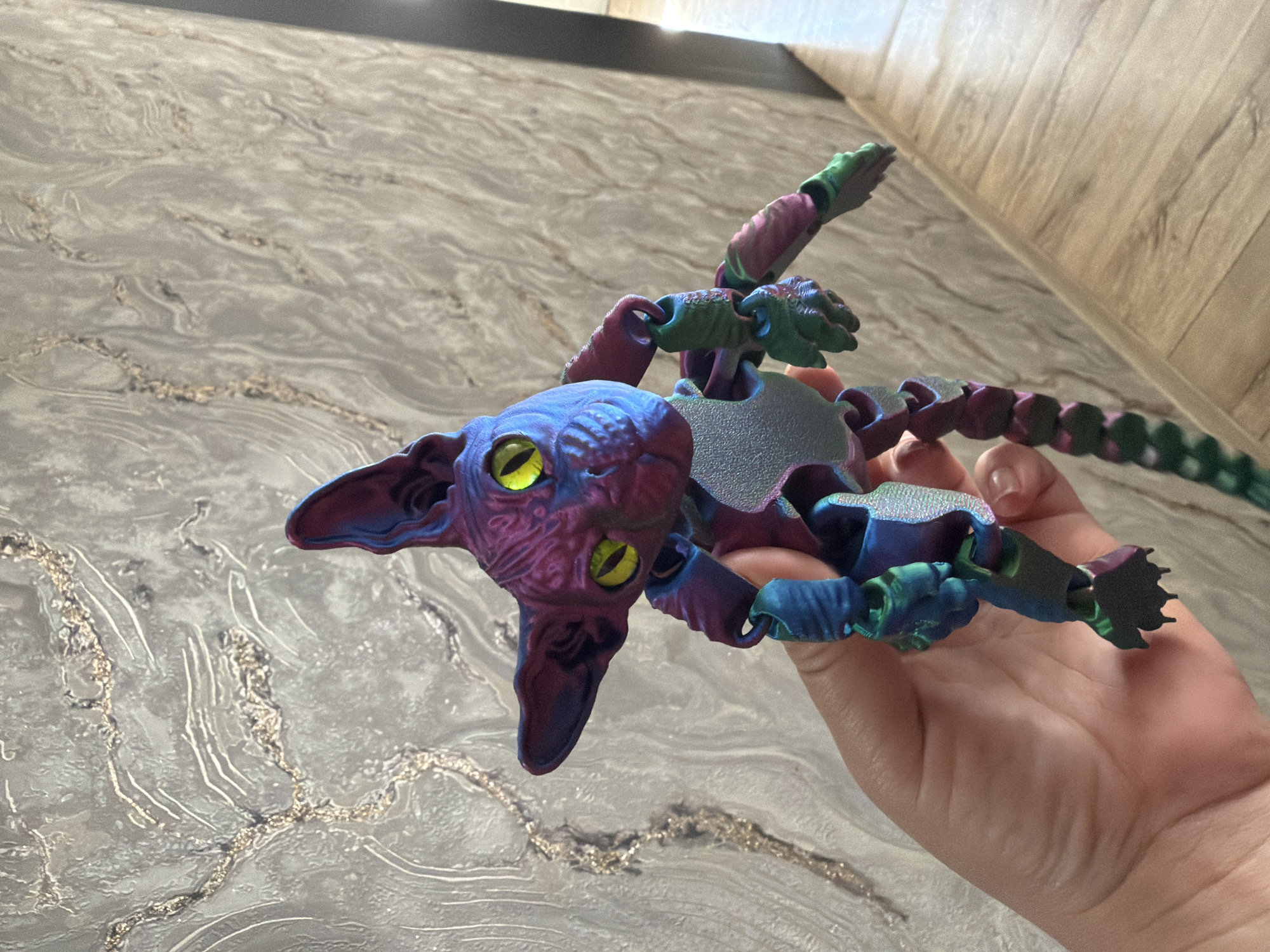 A person's hand holds a 3D-printed articulated toy resembling a Sphynx cat. The toy is multi-colored, with iridescent blues, purples, and greens, and has large yellow eyes. The background consists of a textured wall with a gold metallic pattern and a wooden floor. The scene appears to be indoors, possibly in a residential setting.