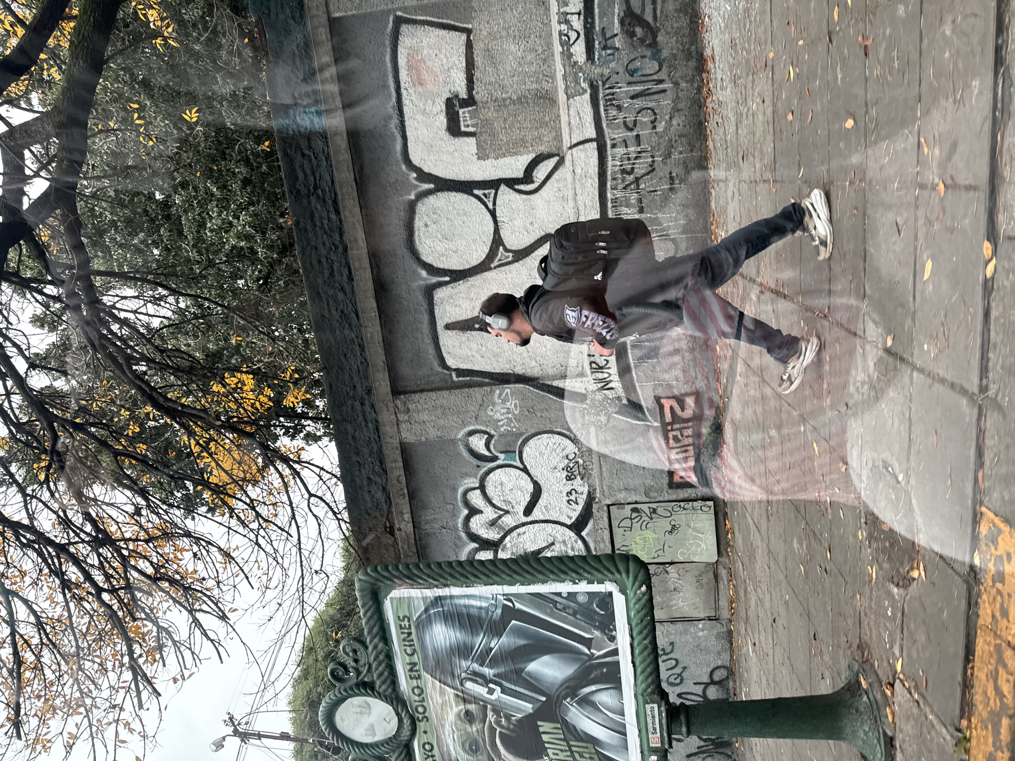 The image, likely taken from inside a vehicle, shows an outdoor scene in Buenos Aires, Argentina. A man with headphones and a backpack is captured mid-stride on the sidewalk, adjacent to a wall covered in graffiti. A promotional poster, framed in green, advertises the movie "The Mandalorian," stating in Spanish "Solo en cines" (Only in theaters). The street sign reads "Sarmiento." Bare tree branches with some yellowing leaves are visible above. The pavement appears wet with fallen leaves scattered across it.