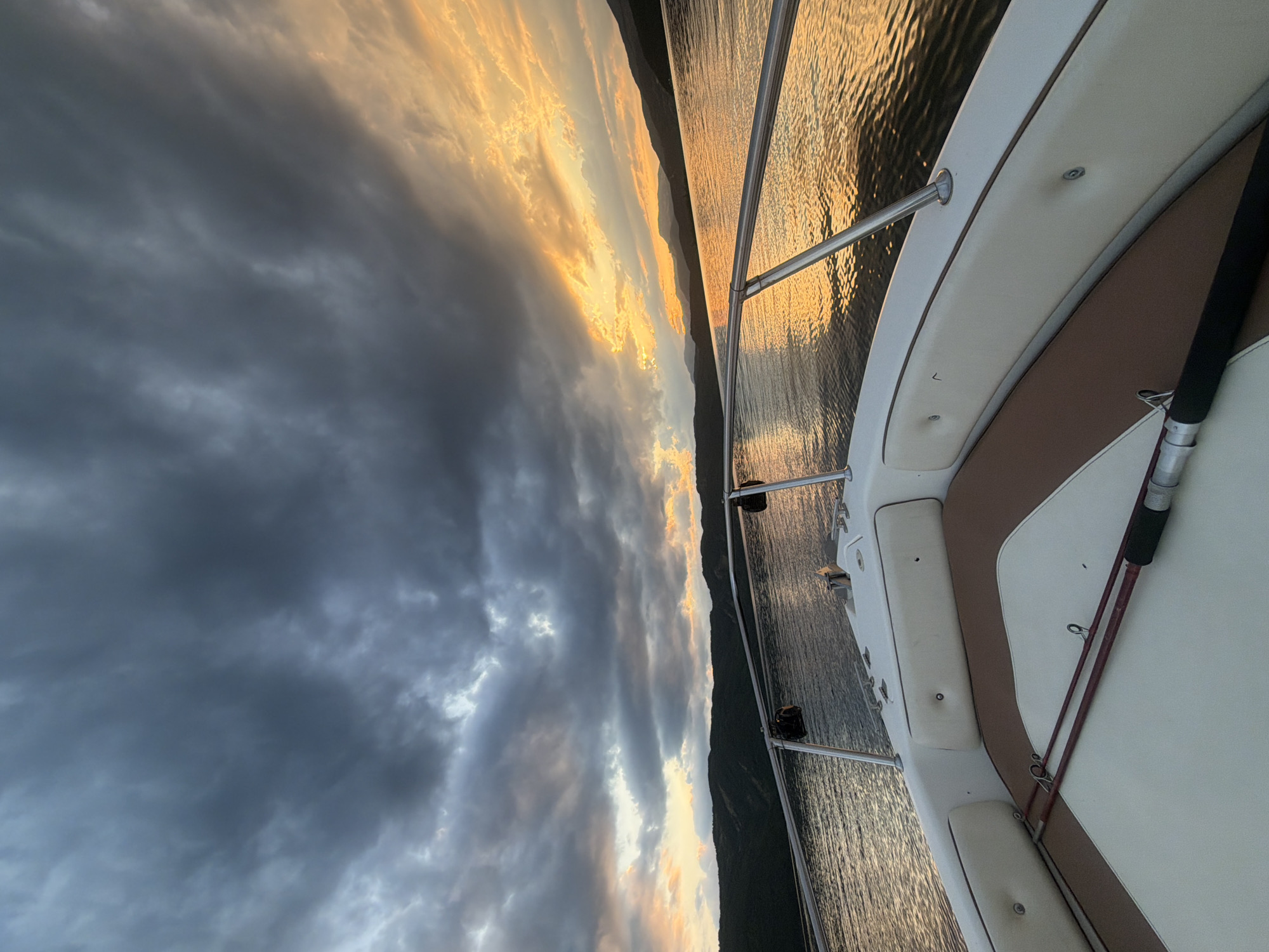 The image captures the bow of a white boat moving across a body of water, likely a lake or reservoir, with a dramatic sunset in the background. Dark, ominous clouds dominate the upper left portion of the sky, contrasting sharply with the vibrant orange and yellow hues of the sunset on the right. The water reflects the colors of the sky, creating a shimmering, golden surface. In the distance, a dark, mountainous shoreline is visible, silhouetted against the bright sky.
On the deck of the boat, a fishing rod with a dark handle and reddish-brown rod lies diagonally across the seats. The seats themselves are upholstered in white with brown trim. A stainless steel railing runs along the sides of the boat. Two black lights or speakers are mounted on the railing.
The scene appears to be set in or near Glavatartsi, Bulgaria, a town known for its reservoir, the "Kardzhali Dam," which is a popular location for water activities and fishing, aligning with the presence of the fishing rod in the image. The overall mood is one of impending weather change, juxtaposed with the beauty of a sunset over a tranquil landscape. There are no people visible in the image.