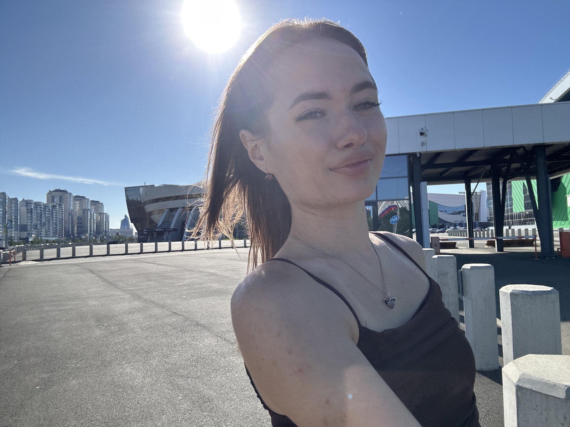 A young woman with brown hair, wearing a dark brown tank top and a silver necklace with a pendant, is taking a selfie outdoors. She has on subtle makeup and is looking towards the camera with a slight smile. The sun is bright, casting a lens flare in the upper left corner of the image. Behind her, there is a large, modern building with glass windows and a dark roof, possibly a stadium or convention center. In the background, there are tall city buildings and a clear blue sky. The foreground shows a paved area with concrete bollards. The overall scene appears to be in an urban environment on a sunny day.