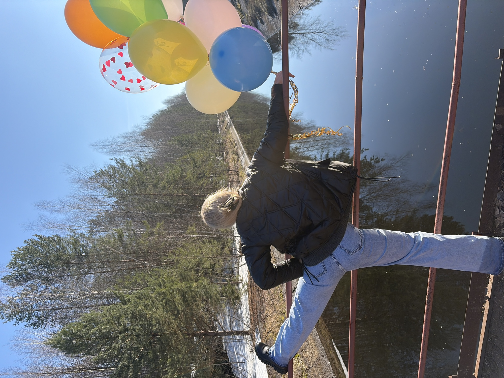 The image depicts a woman hanging from a rusty bridge in Chernushka, Russia. She is holding a bunch of colorful balloons, including orange, green, yellow, blue, and a clear balloon with red hearts. She is wearing a black quilted jacket, light blue jeans, and black shoes. The bridge is over a body of water that appears to be dark and reflective. The background features a landscape of trees and a clear blue sky. Some snow is visible on the ground near the bridge.