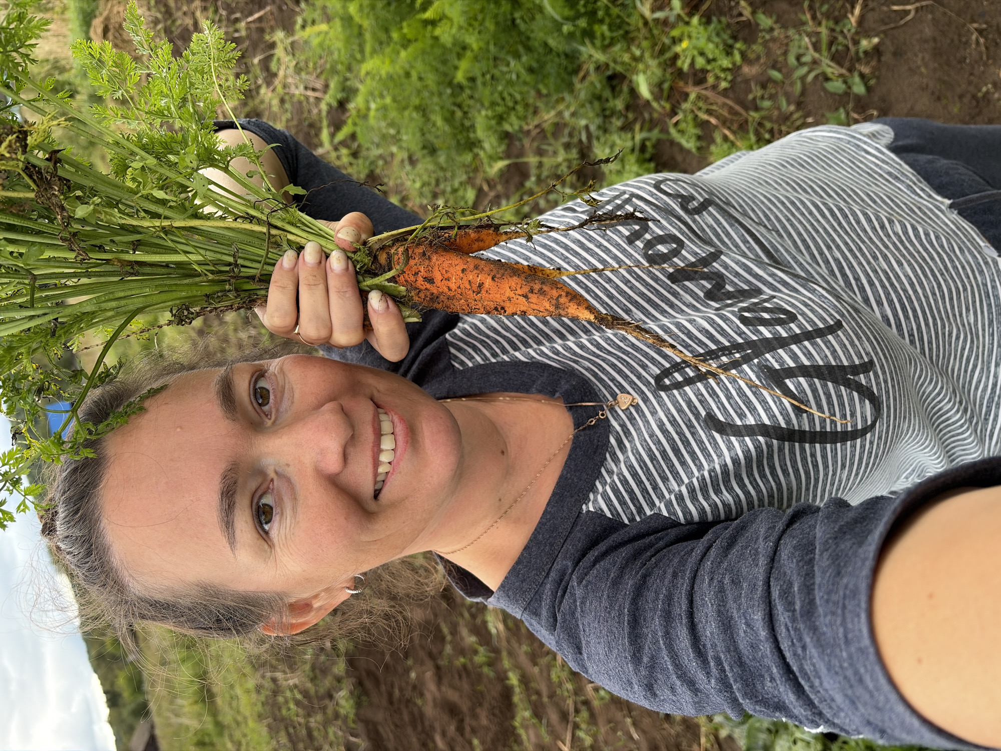 A woman with brown hair pulled back smiles at the camera, holding a bunch of freshly harvested carrots. She is wearing a gray and white striped long-sleeved shirt and dark pants. The background shows a field with rows of dirt and green plants, suggesting an agricultural setting. The sky is overcast. The location is the city of Uzyar, Russia.