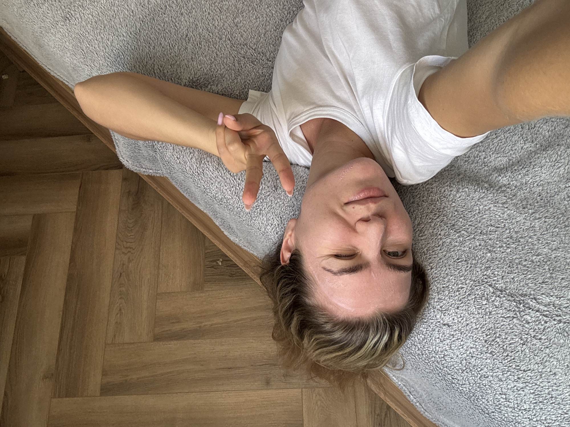 A woman is lying on her side on a gray, textured blanket. She is wearing a white t-shirt. Her left arm is bent at the elbow and her hand is making a peace sign gesture. She is winking and looking at the camera. Her hair is light brown and appears to be pulled back. She has a light application of what appears to be a facial mask on her forehead and cheekbones. The background is a wooden floor with a herringbone pattern. The image is a selfie taken from a high angle. The location is not identifiable.