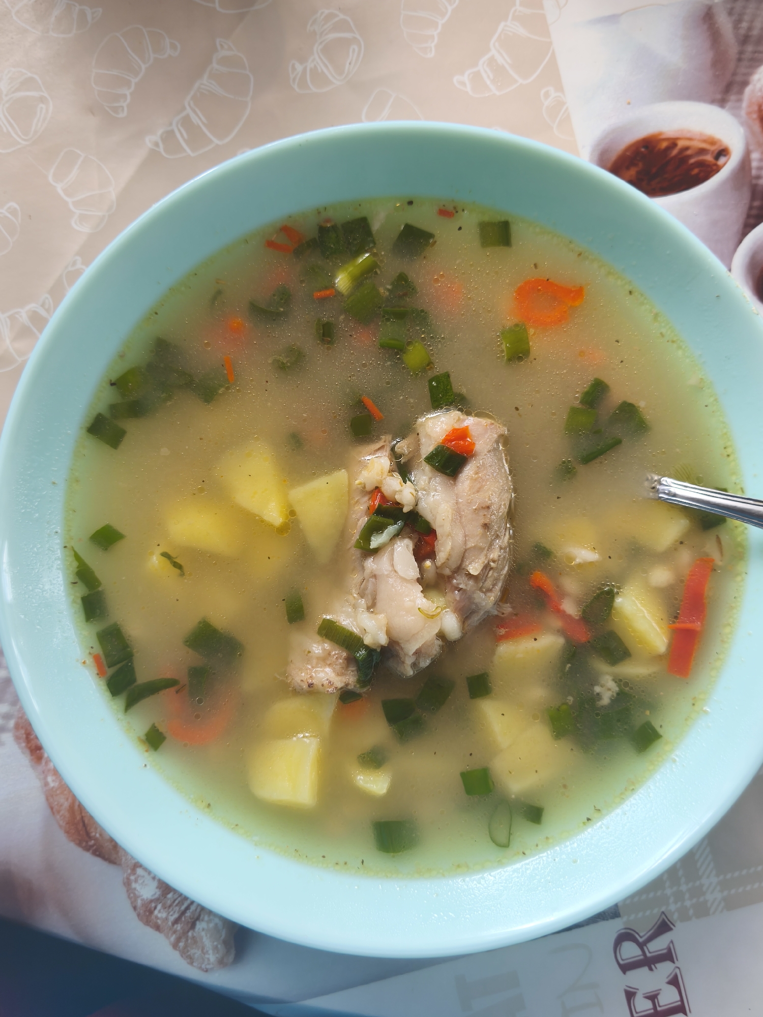 A top-down shot shows a light blue bowl of chicken soup. The soup contains chicken pieces, diced potatoes, carrots, and green onions in a broth. A silver spoon is visible in the soup on the right side of the bowl. The bowl sits on a surface with a pattern of croissants and coffee cups. It is impossible to verify the precise location of this image in the city of Парэчча, Belarus from this image.