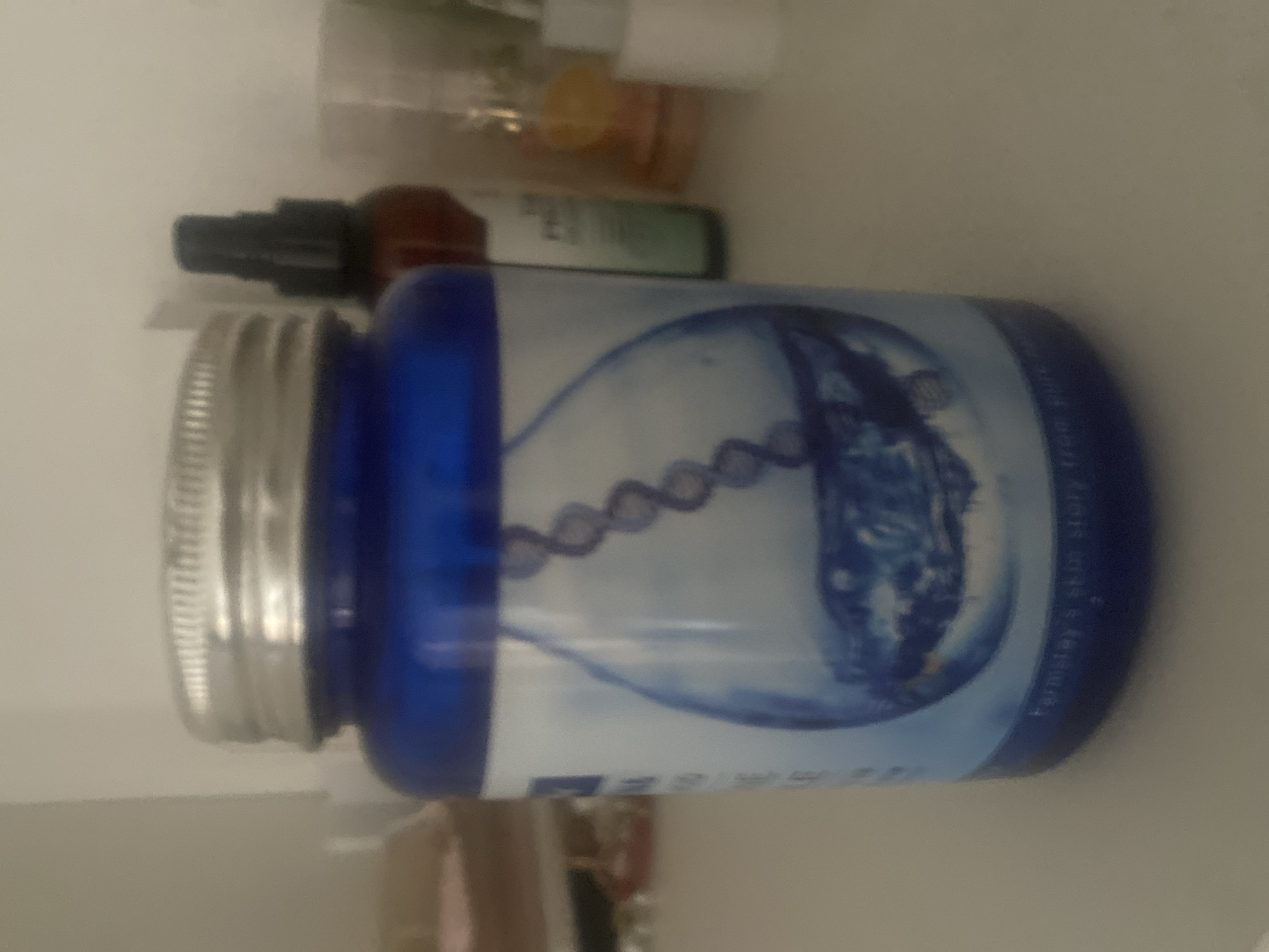 In Boraldai, Kazakhstan, a blue glass jar with a silver lid sits on a white surface, possibly a shelf or table. The jar's label features a prominent image of a water droplet containing a stylized DNA strand and a reflective surface that appears to be water. The text on the label is mostly illegible, though the words "farmacy" and "skin story" are partially visible near the bottom. Behind the jar, other items are visible, including a dark-colored spray bottle and other indistinct containers, suggesting a collection of cosmetic or personal care products. The lighting is dim, and the background is out of focus, creating a soft, indistinct environment.