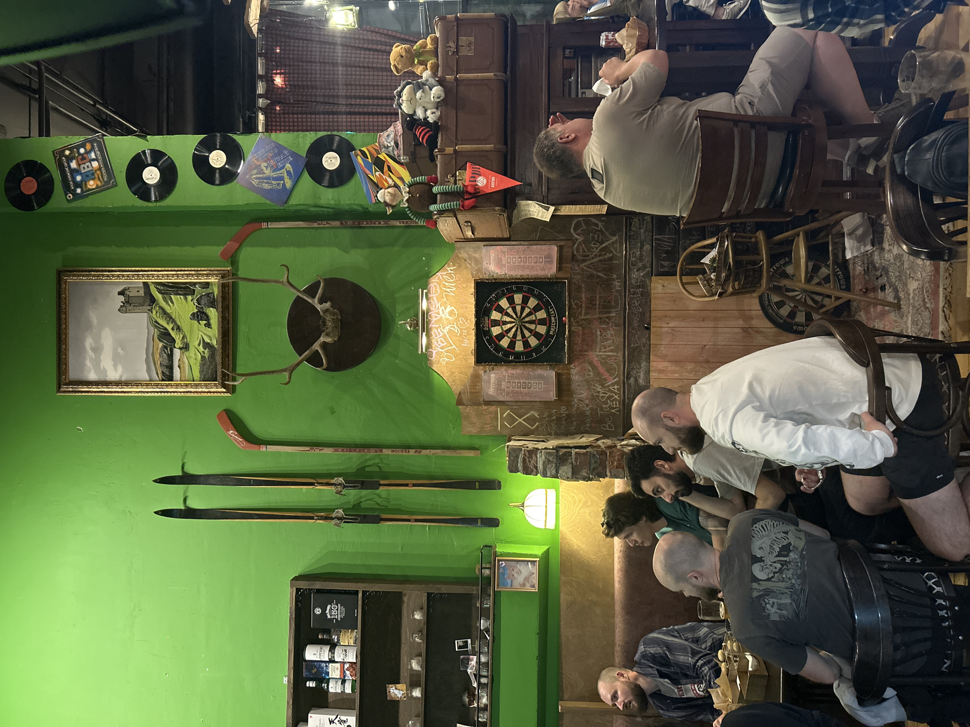 The image depicts an interior scene, likely a bar or restaurant, with a bright green wall as a dominant feature. Several people are seated at tables, some facing away from the camera, engaged in conversation or looking at something off-frame. On the green wall, vintage skis and hockey sticks are mounted, along with a framed painting of a castle and landscape, deer antlers, and several vinyl records. Below these decorations, a dartboard is prominently displayed on a textured wall, with chalk markings and graffiti visible on the surrounding surfaces. The overall atmosphere suggests a casual, possibly retro-themed establishment. The presence of a Russian language sign on a vintage suitcase and the style of the establishment indicate the location is likely Moscow, Russia.