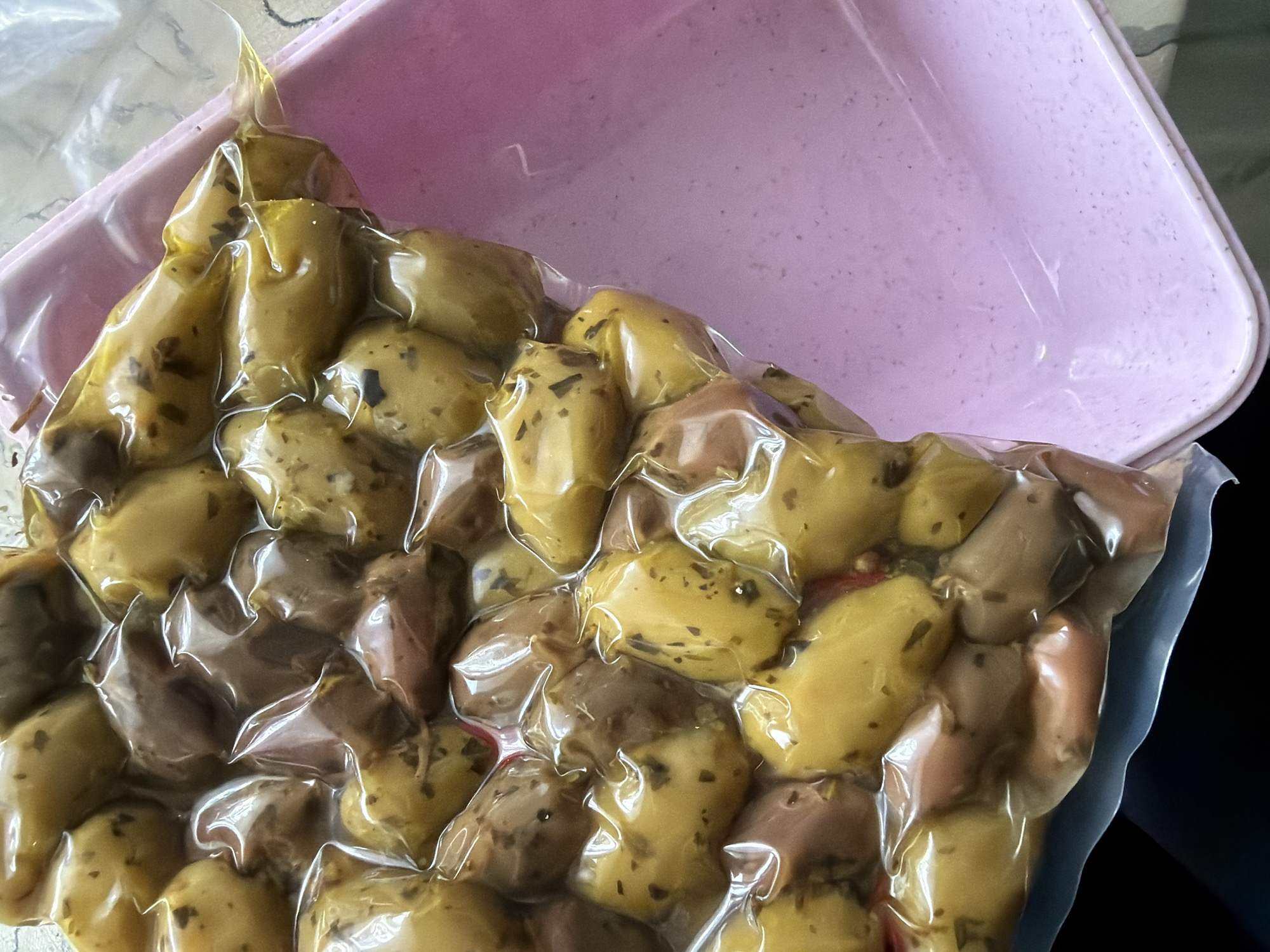 This close-up image displays a vacuum-sealed package of marinated olives, positioned partially within or alongside a light pink plastic container. The olives vary in color, predominantly a yellowish-green hue, with some purplish-brown varieties mixed in. They are coated in a liquid, likely oil or marinade, and speckled with dark green herbs, possibly oregano or thyme. The transparent plastic packaging is tightly sealed, conforming to the irregular shapes of the individual olives and creating reflective surfaces.
The pink plastic container, visible in the upper right, appears empty and features a slightly textured or dusty surface. It provides a clean, contrasting background to the food package. The lighting is bright, highlighting the sheen on the olives and the plastic film. No human activity is depicted, and the scene suggests a food item prepared for sale or storage. There is no visible text on the packaging or container. The setting appears to be an indoor environment, possibly a kitchen or retail display area. No specific cues within the image directly indicate the location as Pazardzhik, Bulgaria, beyond the general context provided.
