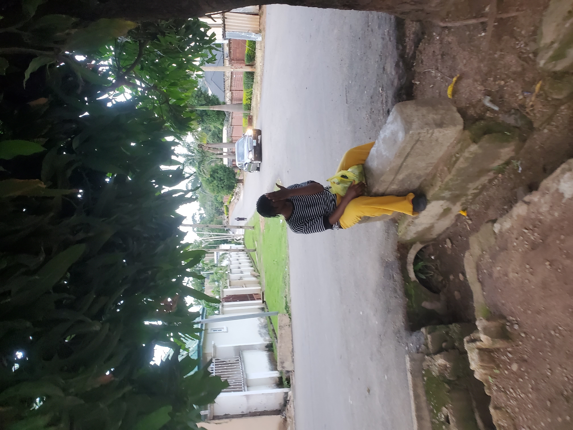 A woman is sitting on a concrete structure beside a road in Jos, Nigeria. She is wearing a black and white striped shirt and yellow pants, and holding a yellow plastic bag. Her hand is covering her face. A car is driving on the road in the background. The scene is partially shaded by trees, and there is some greenery and buildings visible. A drainage ditch is in the foreground.