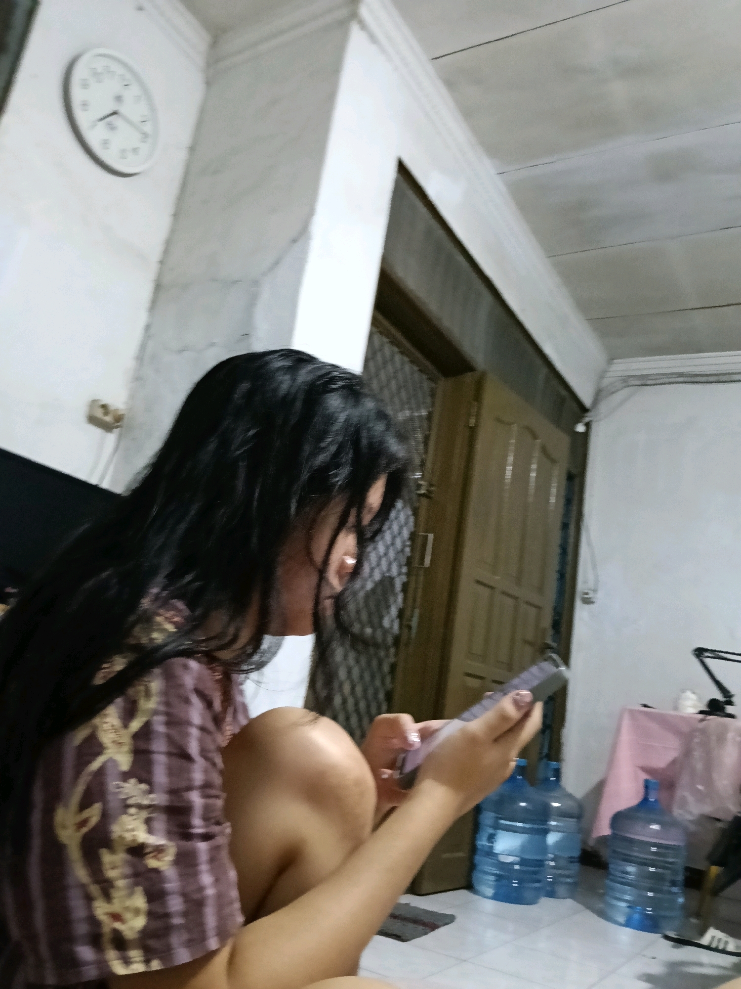 The image shows a young woman with long, dark hair, wearing a patterned shirt, sitting on the floor and looking at a smartphone in her hands. She is indoors, in what appears to be a room. In the background, there is a white wall with a clock on it, a door, and several large water bottles. The room has a tiled floor and the ceiling is visible. The lighting suggests it may be daytime, or an indoor light source is on. The overall scene is casual and domestic.