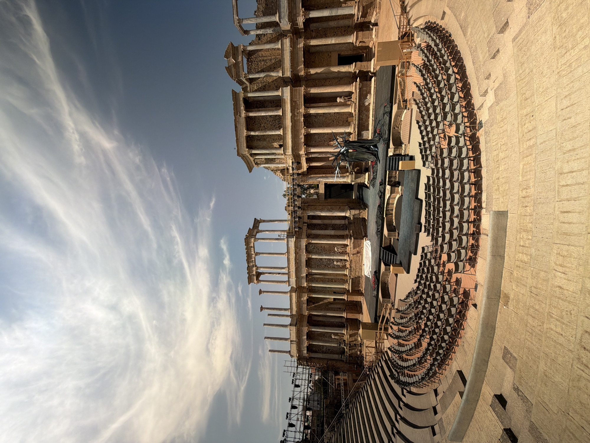 The image depicts the Roman Theatre of Merida, Spain, an ancient amphitheater still used for performances. The sun is shining under a sky with wispy white clouds. In the foreground, the tiered seating area is empty and made of stone. Rows of dark chairs are set up on a circular section of the stone seating.
The stage area is visible in the middle ground. It is a dark, raised platform with a backdrop of stone ruins featuring columns and arches. A figure dressed in black, appearing to be an actor in costume, stands center stage next to a dark, tree-like prop decorated with red fabric. Scaffolding and lighting equipment are visible to the left of the theatre ruins. The overall scene suggests a preparation for a performance in a historical setting.