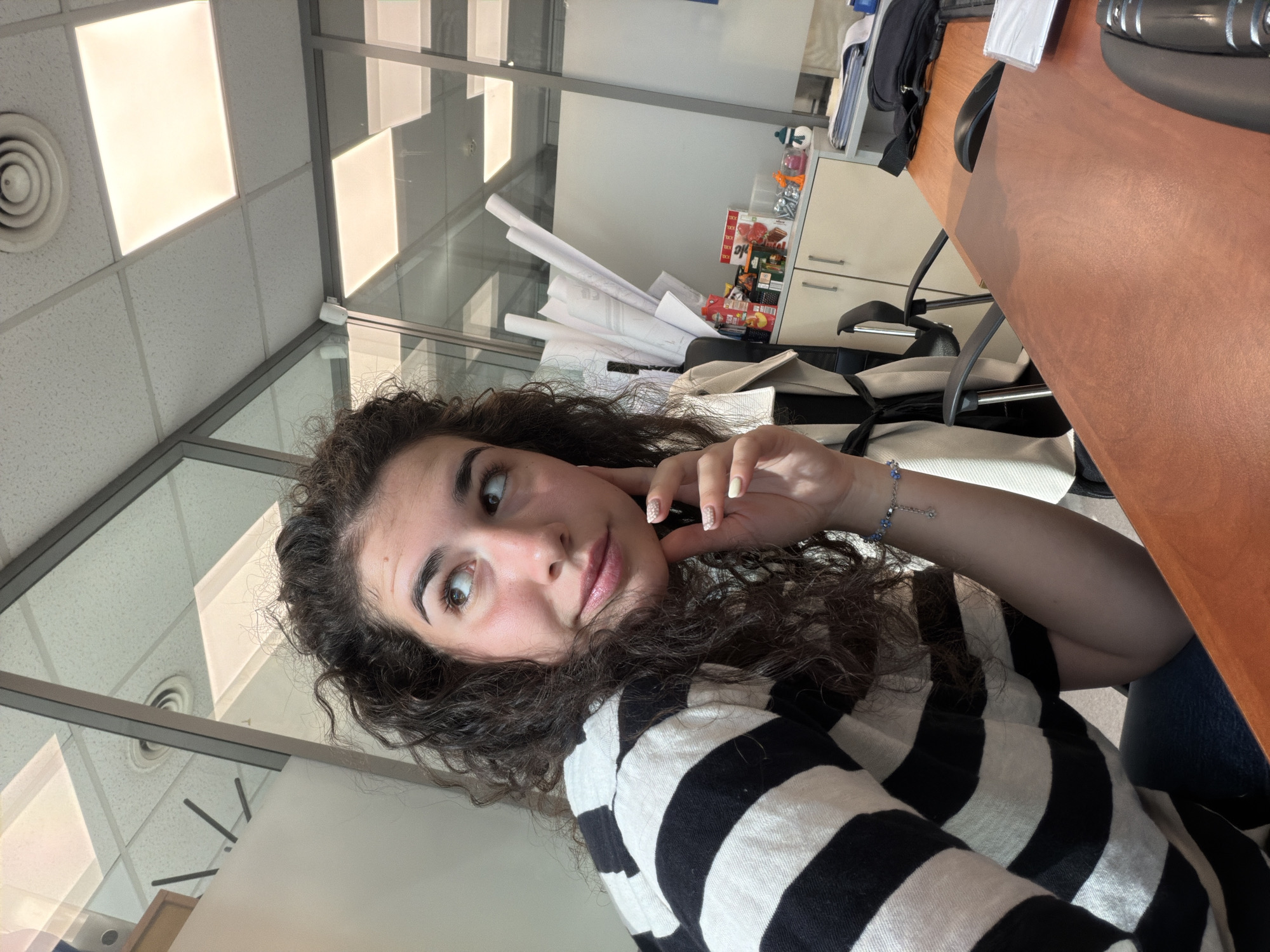 A young woman with curly dark hair is sitting at a wooden desk in an office. She is wearing a black and white striped shirt and looking up and to the left with her hand on her chin, her expression suggesting thoughtfulness or contemplation. Her fingernails are painted with a light color and have small dot designs. A bracelet with small blue charms is visible on her wrist. The office environment appears modern, with glass partitions and ceiling lights. In the background, there are office cabinets with various items on top, including rolled-up papers, what appears to be a juice carton, and other small objects. A computer mouse and keyboard are visible on the desk. The scene is indoors, and the lighting suggests it is daytime. The image does not contain any information to place it in Sofia, Bulgaria.
