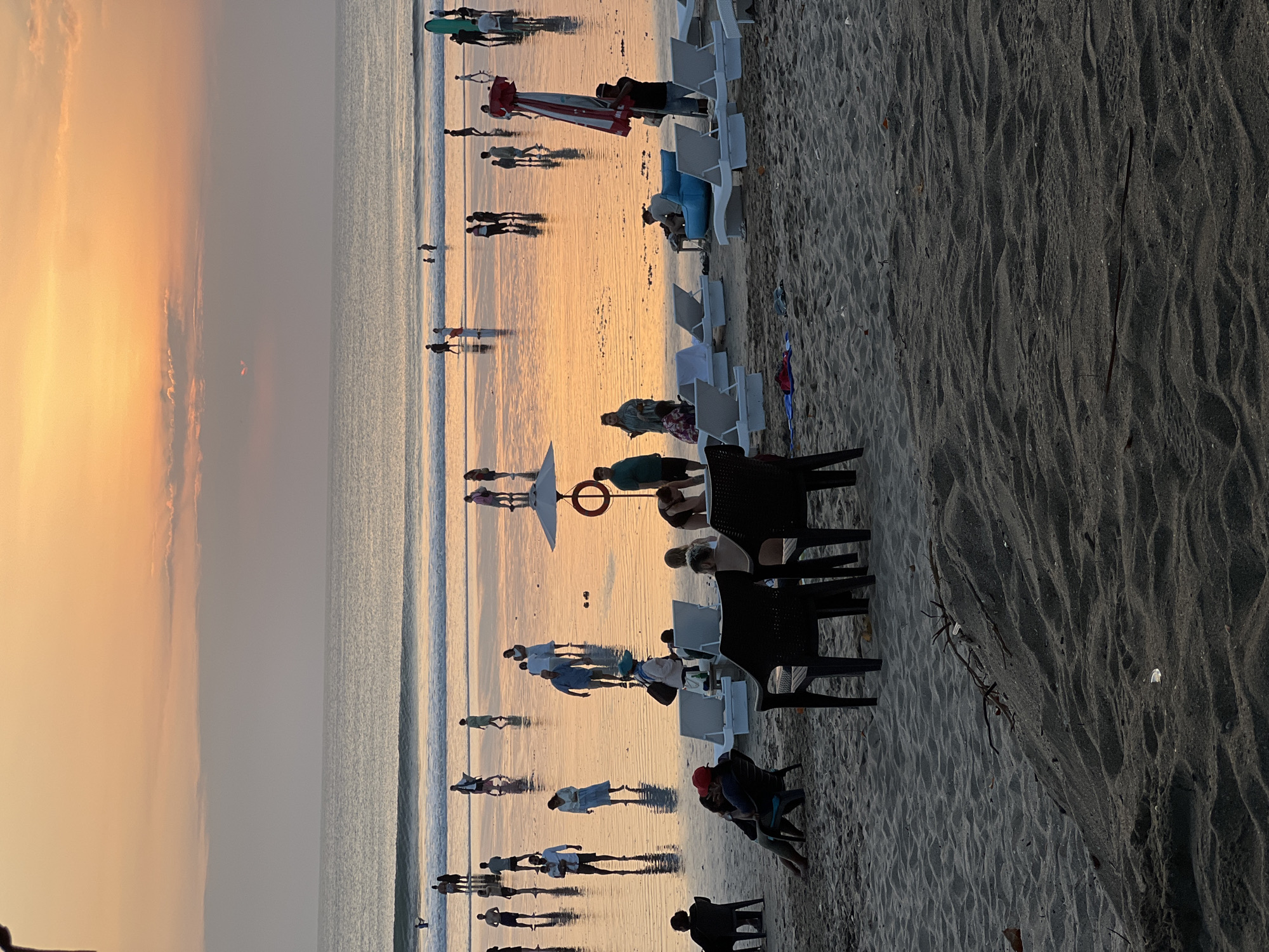 This photograph captures a serene beach scene at sunset in Kuta, Bali, Indonesia. The sky is painted with soft hues of orange and pink, transitioning into a pale yellow at the horizon. The ocean reflects the warm colors of the sunset, creating a shimmering effect on the wet sand.
Numerous people are scattered across the beach. Some are walking along the water's edge, while others are standing or sitting further back on the sand. Several individuals are wading in the shallow water, enjoying the last light of the day.
In the foreground, there are several beach chairs and lounge chairs, some of which are occupied. Two black plastic chairs are positioned in the center foreground, with a couple seated in one. Further back, white lounge chairs are arranged, some facing the ocean, creating a sense of relaxation and leisure.
Objects like a life preserver on a pole, a surf board leaning against a stand, and an umbrella are visible, suggesting a typical beach setting. The sand itself is a prominent element, with textured patterns from the ebb and flow of the tide and footprints from the beachgoers. The overall atmosphere is one of tranquility and natural beauty, enhanced by the warm glow of the setting sun.