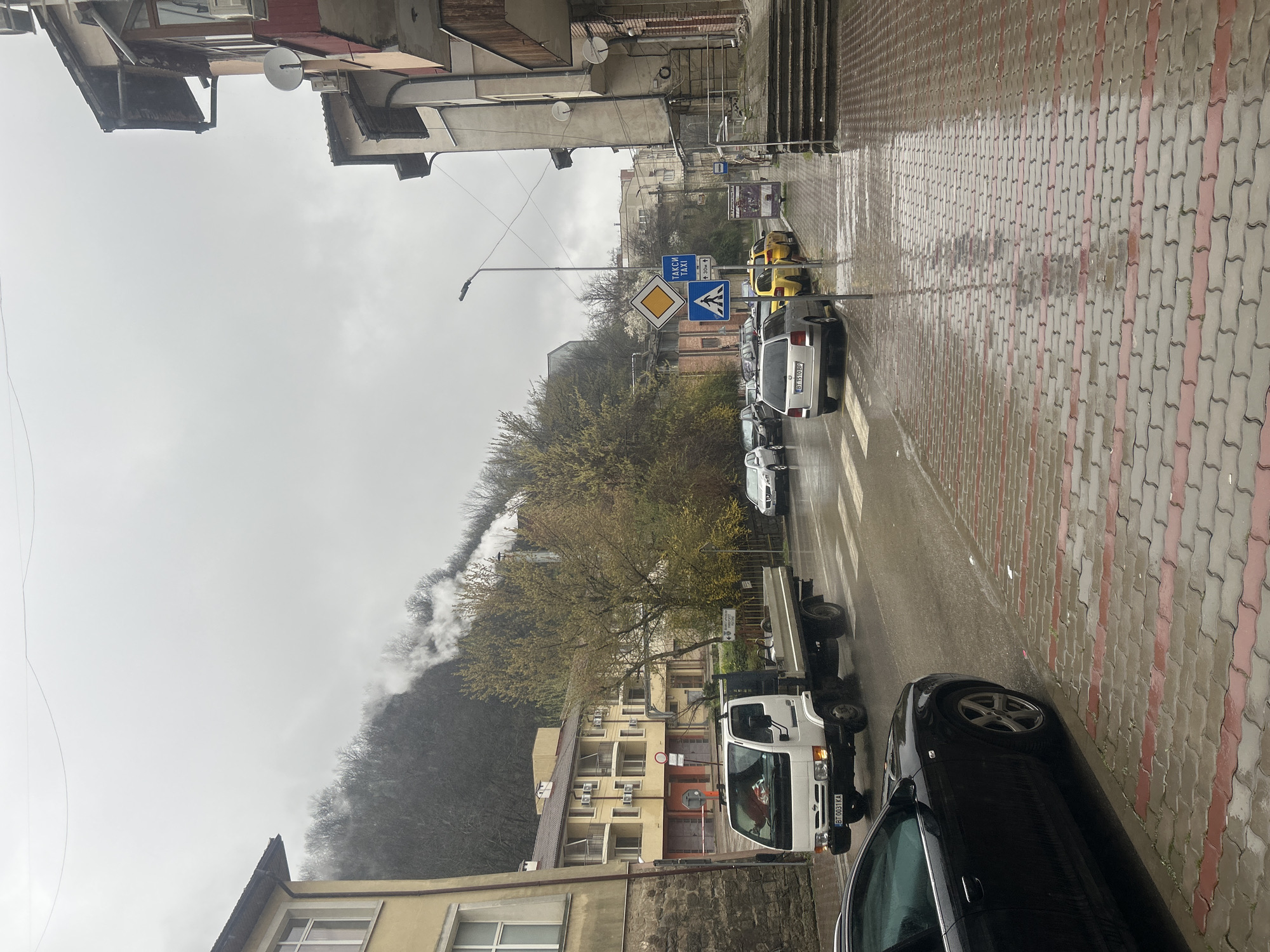 The image depicts a wet, overcast street scene in Veliko Tarnovo, Bulgaria. The street is lined with buildings, a few of which appear to be older. Vehicles are parked along the right side of the street, including a white truck, a silver SUV, and several other cars. The street is paved with interlocking bricks, and the sidewalk is also paved with bricks of a similar pattern. There are several road signs visible, including a pedestrian crossing sign and a sign indicating a taxi stand. In the background, a tree-covered hill is partially obscured by fog or clouds. The overall impression is of a quiet, rainy day in a small town.