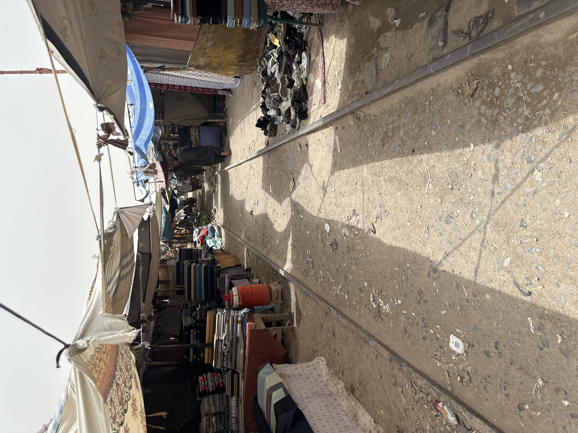 The image depicts a bustling marketplace in Kuchlak, Pakistan, with a central pathway lined by vendors. The pathway appears to be an abandoned railway track, evidenced by the two parallel metal rails running down the center. The market stalls are covered with various awnings, some made of patterned fabric and others of blue tarp. On the left side, stacks of colorful textiles and fabric rolls are neatly arranged for sale. A large orange cooler is placed on a stool next to the fabric stalls. On the right side of the pathway, a large pile of assorted footwear, including sneakers and sandals, is displayed on a rug. Several individuals are visible in the background, appearing to be shoppers and vendors. The ground is a mix of dirt and gravel, littered with debris. The overall scene suggests a vibrant commercial activity taking place under a bright, overcast sky.