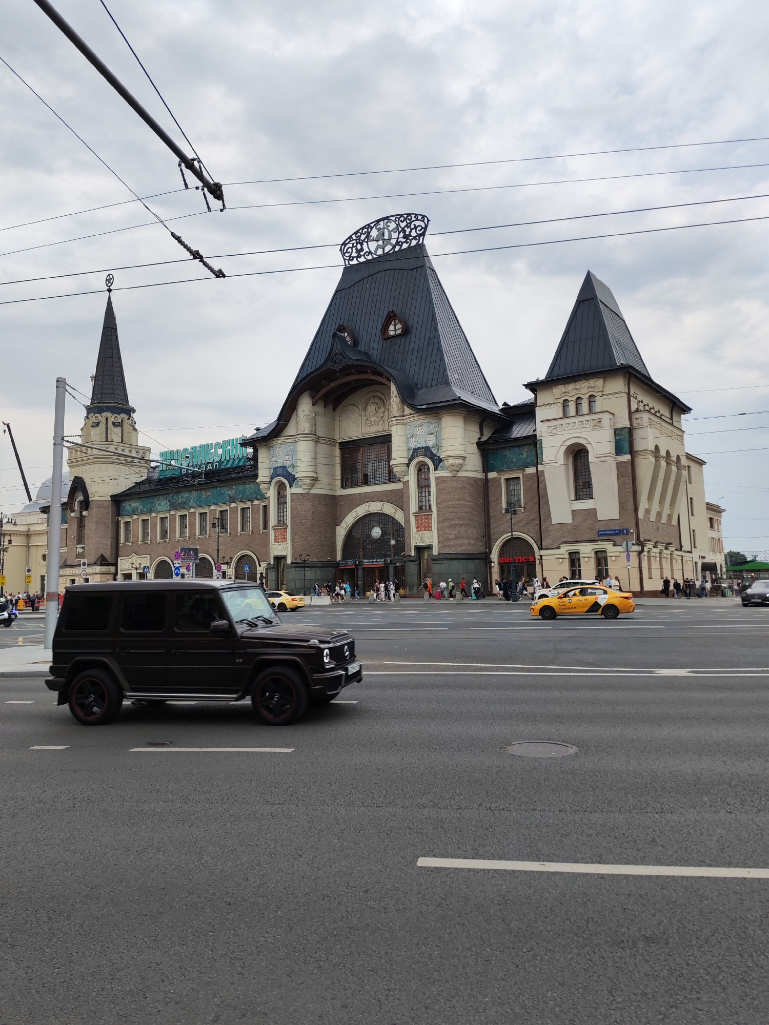 This is a photograph of a busy street scene in Moscow, Russia, likely in front of a train station. The most prominent structure is a large, ornate building with multiple spires and towers, characteristic of Moscow's historic architecture, possibly the Yaroslavsky Rail Terminal given the signage on the building. In the foreground, a dark-colored SUV is driving on the asphalt road. Further down the road, a yellow taxi cab is also visible, along with several pedestrians walking near the building's entrance. Overhead, electrical wires for public transportation are strung across the sky, and the sky is overcast with clouds. The overall impression is that of an active urban environment on a cloudy day.
