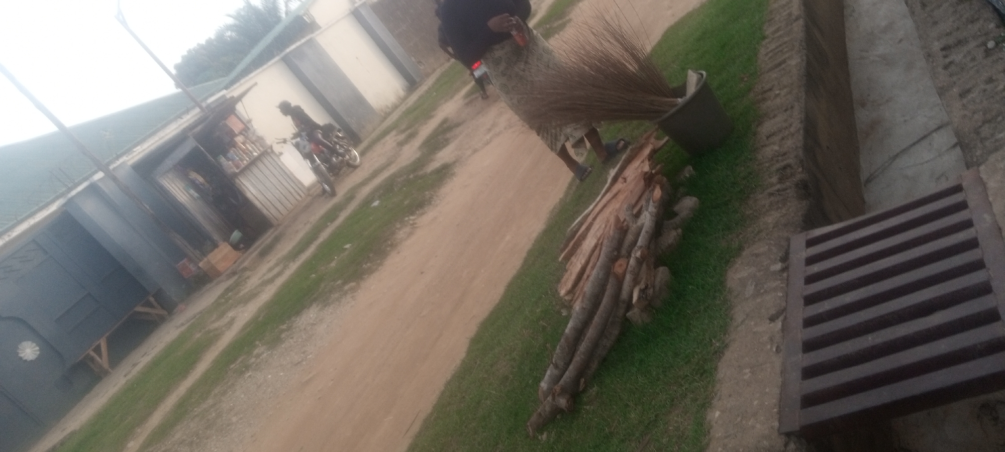 The scene appears to be an outdoor location in Narayi, Nigeria. A dirt road runs through the center of the image. A building, possibly a shop, stands on the left side of the road. Parked near the shop is a motorcycle with one person on it. On the right side of the road, firewood is stacked on the grass. A woman in a long skirt stands near a bucket containing brooms. A storm drain is located in the lower right corner of the image.