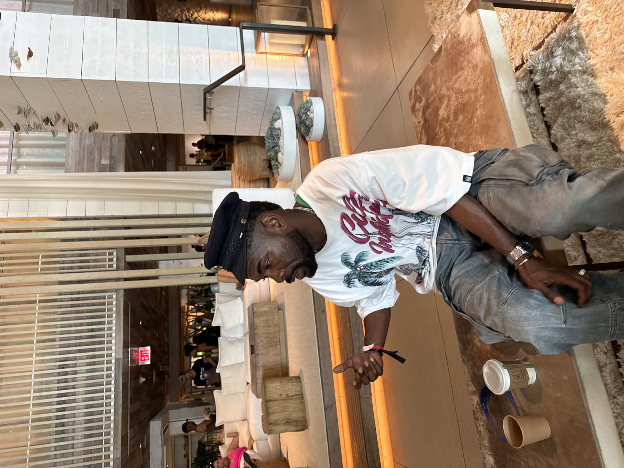 This media file captures an indoor scene, likely a modern hotel lobby or lounge, in Miami Beach, United States of America. The image is oriented vertically.
The main subject is a dark-skinned man with a beard, positioned to the right of the frame. He is wearing a black flat-top hat with a metallic badge, a white t-shirt featuring a graphic that includes palm trees and red stylized text reading "Miami," light-colored denim jeans, a silver-toned watch on his left wrist, a ring on his left ring finger, and a red wristband on his right wrist. He appears to be seated or leaning, looking downwards and to his left, with his right hand extended as if gesturing or pointing, and his left hand resting on his thigh.
The background reveals a spacious, well-lit interior. To the left, white modular sofas with numerous white cushions and wooden block-like tables or stools suggest a lounge area. Further back, more people are visible, seated and appearing to socialize. Decorative elements include white slatted or paneled structures, some adorned with hanging leaf-like or butterfly-like ornaments, and low planters containing succulents. The flooring consists of light-toned tiles and a shaggy brown rug in the foreground. A red "EXIT" sign is visible on a distant wall.
On the floor near the man's right foot, there is a white disposable coffee cup with a lid, featuring a green and white logo, and a beige paper cup, along with a blue strap or handle. The artificial lighting suggests it could be evening or a space designed to rely primarily on indoor illumination. The overall ambiance is relaxed and social.
