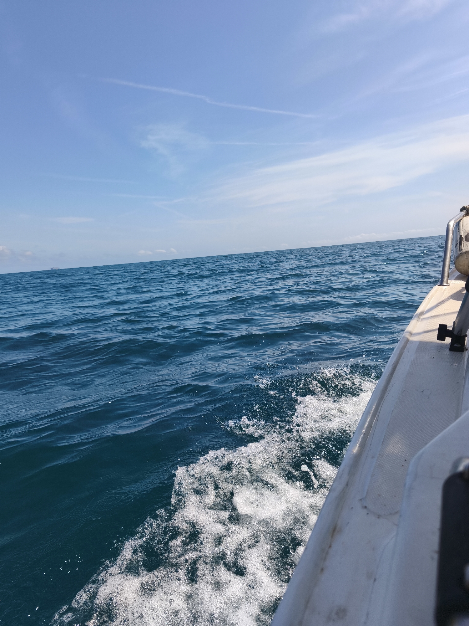 A forensic analysis of the image depicts a daytime scene on open waters off the city of Sueca, Spain. The perspective is from the starboard side of a moving white boat, with its hull and a section of a metallic railing or support visible in the right foreground. The boat generates a substantial white wake and foam along its side as it traverses the deep blue, moderately choppy water. The sea extends to a clear horizon line, where a large, dark, indistinct object, possibly a distant ship or offshore structure, is faintly visible to the left of center. The sky above is a light blue, featuring sparse, wispy white clouds and several faint contrails. No individuals are present in the frame.