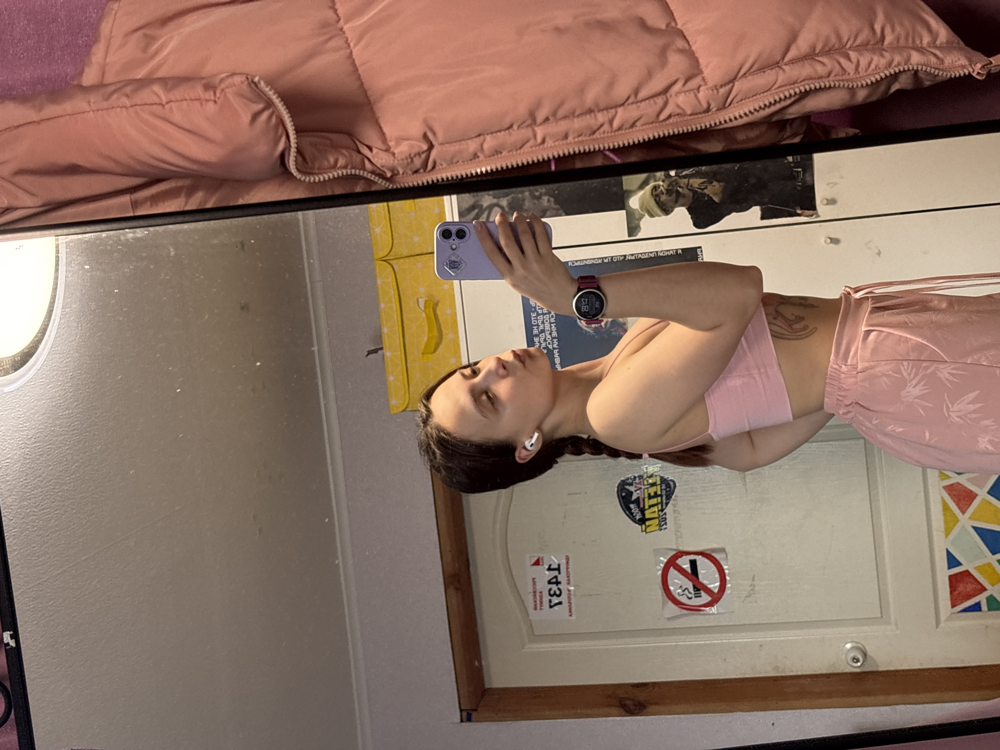 The image shows a person reflected in a mirror. The person is holding a smartphone to take a photo. She is wearing a pink bra top and pink floral print bottoms. She has long brown hair pulled back in a braid and is wearing a watch on her left wrist. The mirror's background reveals part of a room with a pink puffy jacket at the top. On the white door reflected in the mirror, there are stickers and signs including a "no smoking" symbol and a sign with the text "丁431" and "TYMNEA БОССКУСИИ" written on it. There is also text written in reverse, which reads "ARNAYIRO RANХЭРЧУМДУ" and "АНЕ ЭН OTE-D IP UBAL TOAL HBAR AH AHM RD ΟΤΗ ΝΙΗΤΑΔΕΝΗ ΝΟΜΑΤ Α." The sign suggests that the location is in Прикамский, Russia.
