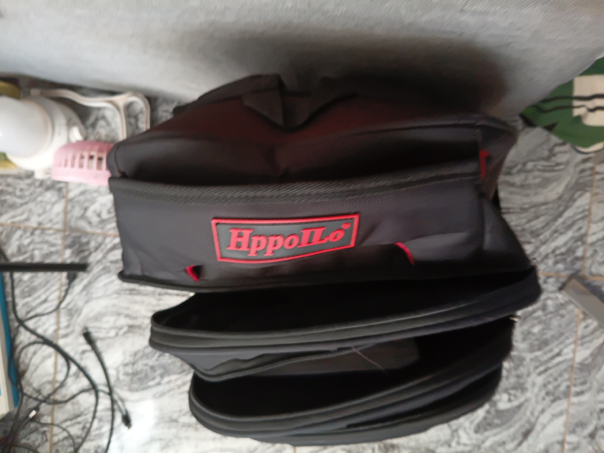 In Jalingo, Nigeria, a black backpack with a red "Hippollo" logo is displayed on a tiled floor with a marble-like pattern. The backpack is partially open, revealing multiple compartments. To the left of the backpack, a black power cord is visible. In the background, a white lamp and a pink object are present, along with a greyish-blue surface. The scene is lit by natural light.