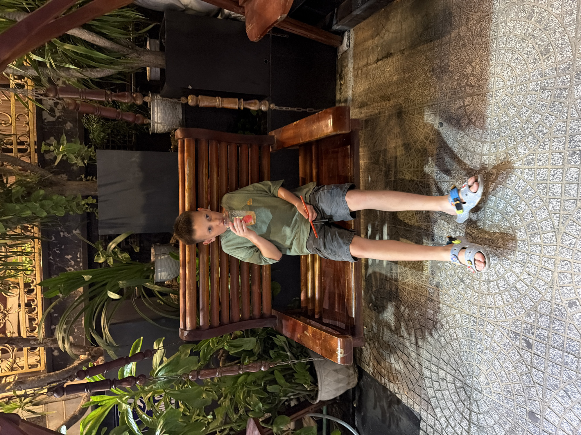 A young boy with brown hair is sitting on a wooden swing bench, holding a clear plastic cup with a straw in his right hand and drinking. He is wearing a green t-shirt and dark gray shorts, with colorful sandals on his feet. The swing bench is positioned on a patterned tile floor, with shadows cast by nearby plants. In the background, there are lush green plants in pots and decorative metalwork. The scene appears to be an outdoor seating area, likely a cafe or restaurant, in the city of Đà Nẵng, Vietnam.
