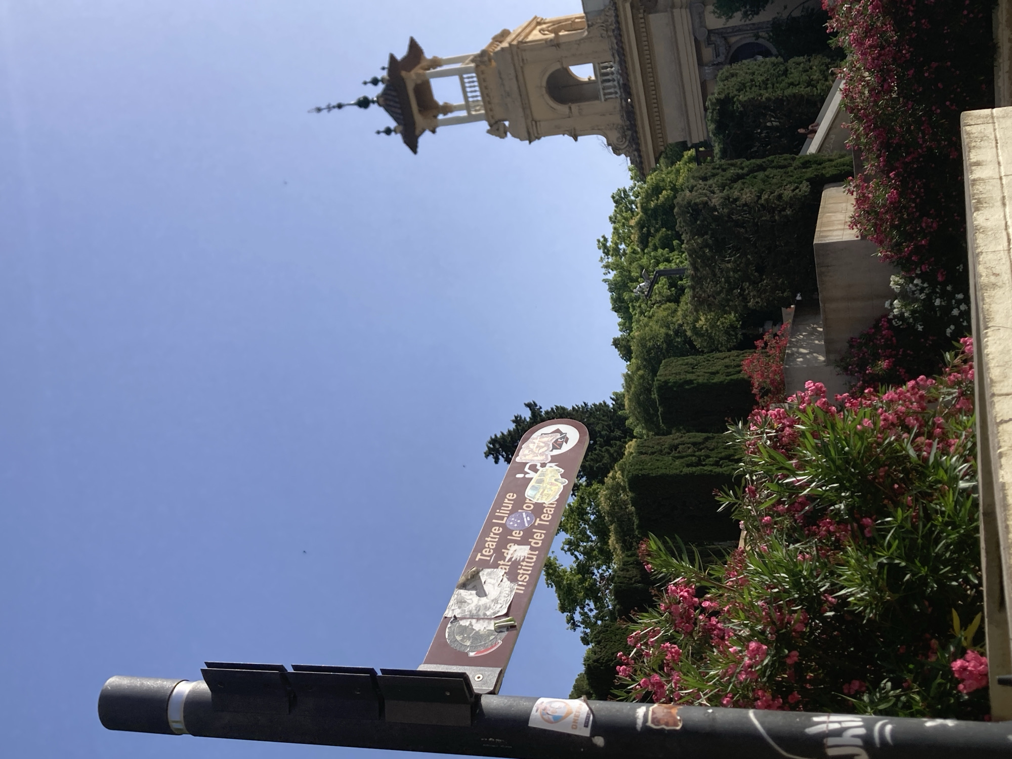 In the city of Barcelona, Spain, this image captures a clear blue sky over a park-like setting. A black lamppost stands prominently in the foreground on the left, adorned with stickers and a brown directional sign. The sign points to the right and reads, "Teatre Lliure / Institut del Teatre," indicating the location of a theater and an institute. In the background, a light-colored tower with a multi-tiered roof and decorative elements stands against the sky. Lush green trees and vibrant pink flowering bushes fill the midground and foreground, creating a colorful and natural ambiance. The scene appears to be a public space, possibly a garden or promenade, with architectural elements suggesting a cultural or historical district.