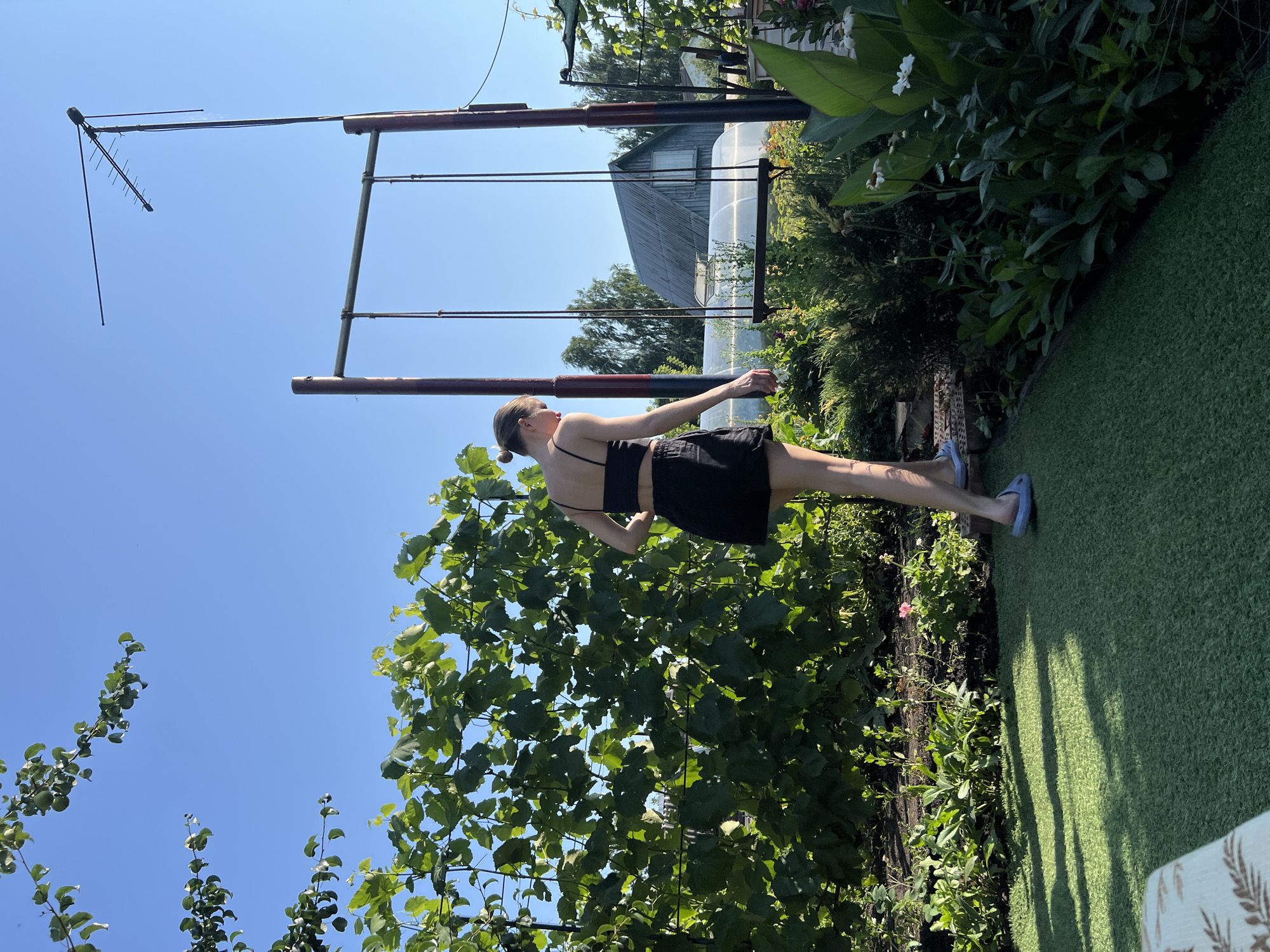 In Lopatino, Russia, on a sunny day with a clear blue sky, a young woman with her hair pulled back in a bun walks on artificial turf in a garden. She is wearing a black crop top and black shorts, and blue slip-on shoes. To her left, a lush green vine-covered trellis runs horizontally across the frame. In the background, a swing set with a wooden seat and dark poles stands under a tall antenna mast. Beyond the swing, a rustic wooden house with a steep roof and a small window is visible, along with a greenhouse structure. The garden features a variety of green plants, bushes, and some white flowers, with shadows cast on the turf.