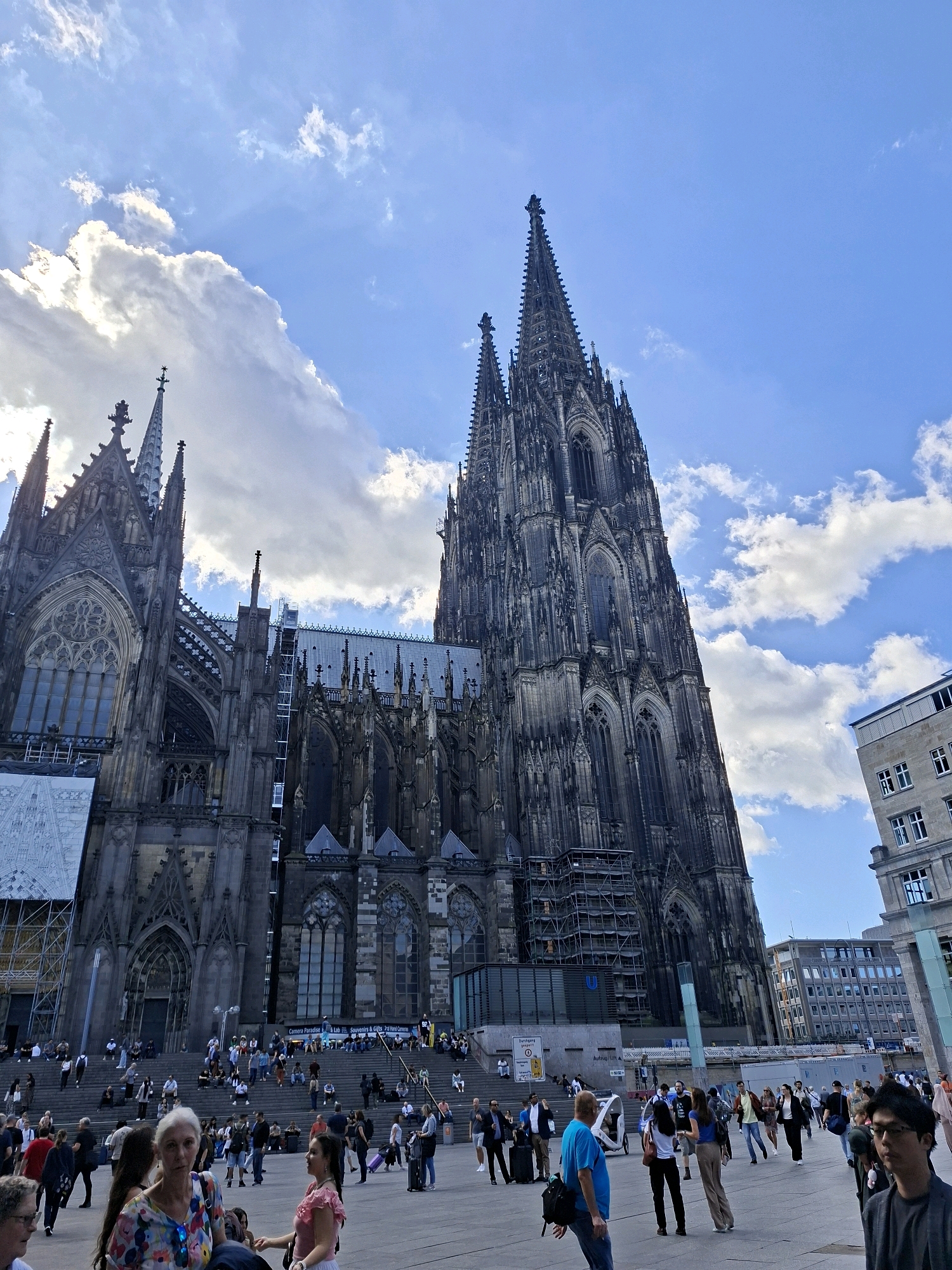 The image captures a wide-angle view of the Cologne Cathedral (Kölner Dom) in Cologne, Germany, on a partly cloudy day. The imposing Gothic cathedral dominates the frame with its intricate spires and detailed architecture. In the foreground, a plaza is populated with numerous people walking, standing, and sitting on the wide steps leading up to the cathedral. Several individuals are in focus, including a woman in the front with a colorful shirt, and a man in a blue t-shirt walking away from the camera. Other people are seen with luggage, suggesting travel. Modern buildings are visible to the right of the cathedral, indicating the urban setting. The sky is a mix of blue with large, fluffy white clouds. Construction scaffolding is visible on parts of the cathedral.