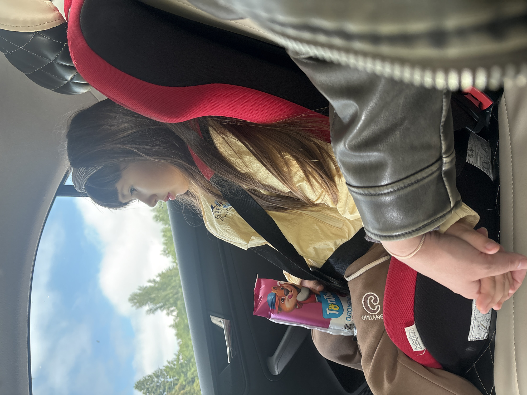 Inside a car, a young girl with long brown hair, wearing a yellow long-sleeved top and brown pants, is seated in a red and black child safety seat. She is holding a pink package with a cartoon fox or wolf character on it, along with the Russian text "TonТоп" and "ПЛОМБИР" (meaning "Plombir" ice cream). She appears to be looking down at the package. Her yellow top has a small cartoon emblem, and her brown pants feature "CHUANG" and a 'C' logo.
In the foreground, an adult's arm, clad in a grey-brown leather jacket and wearing a thin bracelet, is gently holding the girl's hand. A "babyton" label is visible on the child's car seat. Through the car window, a bright, cloudy sky and green trees are visible, indicating it is daytime and the vehicle is likely in motion, possibly outside a city. The overall scene suggests a comforting moment during a car journey in Chaikovsky, Russia.