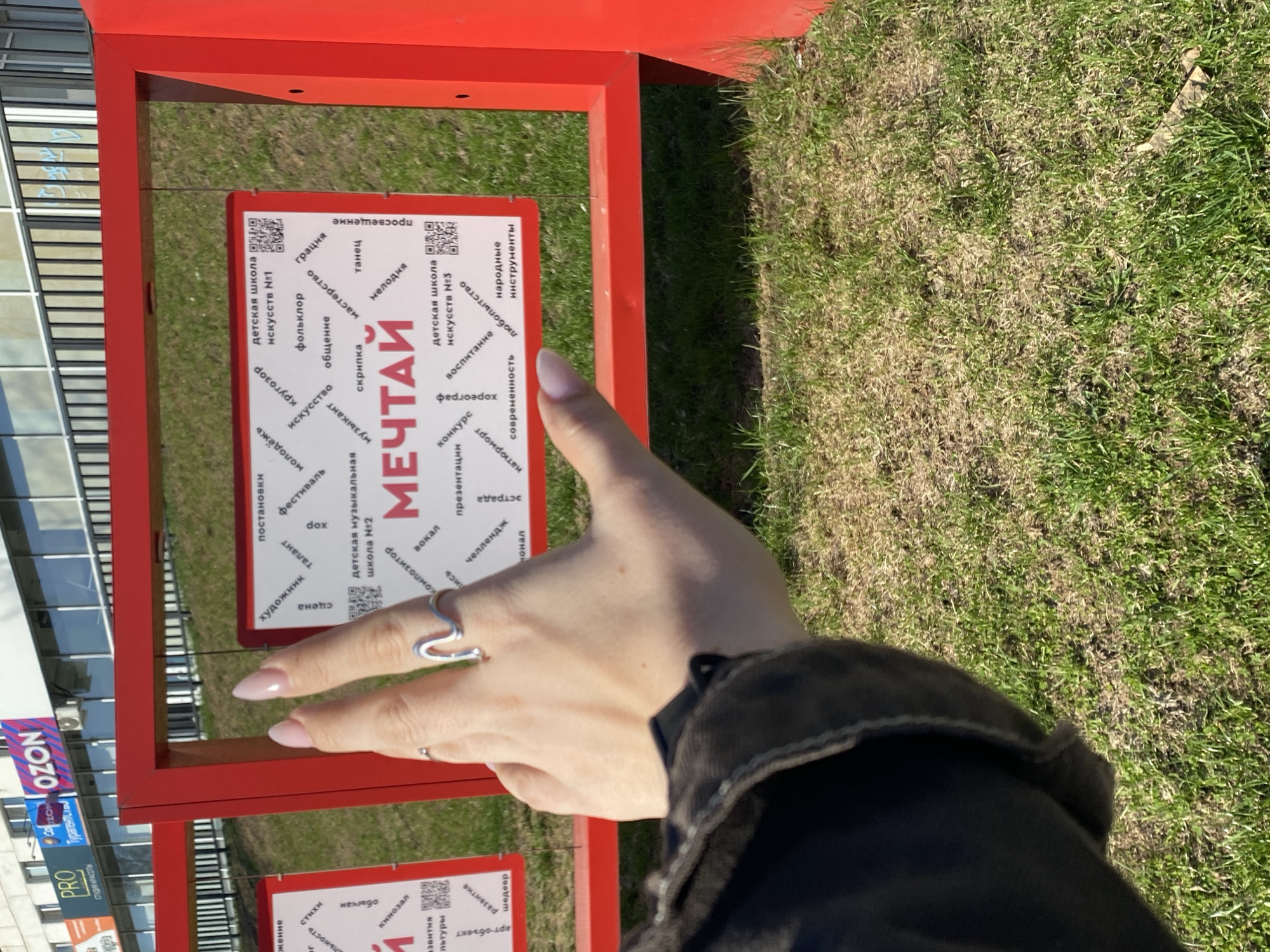 The image shows a person's hand with two silver rings holding up two fingers, set against the backdrop of an outdoor display in what appears to be a park or grassy area. The display consists of red frames each containing a sign. One sign prominently features the word "МЕЧТАЙ" ("DREAM") in Cyrillic script and other related words. The signs also mention детская школа искусств (children's art schools) numbered 1 and 3, and детская музыкальная школа №2 (children's music school number 2). This scene is located in the city of Chaikovsky, Russia. Other words and phrases on the sign include "folklor," "iskusstvo" (art), "obschenie" (communication), and "narodnye instrumenty" (folk instruments). The image captures a building in the background and the advertisement of PROMOZON, a store.