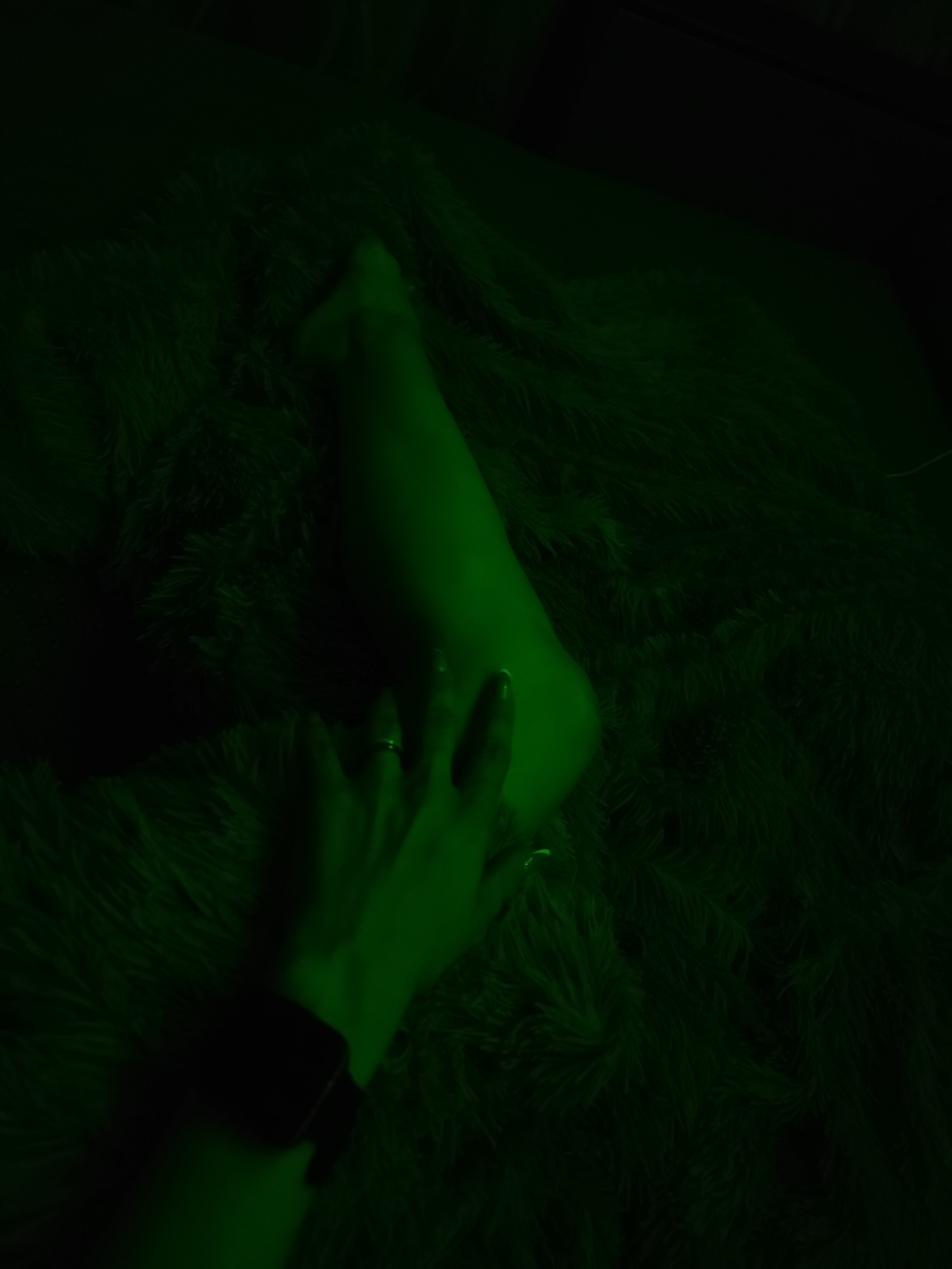 The image shows a green-toned scene with a person's leg resting on a fuzzy surface, possibly a blanket or rug. A hand, also green-toned, is placed near the leg. The person's wrist has a dark object attached, possibly a watch. The details in the scene are obscured by the green lighting. No other information is available to determine a location in the city of Фоки, Russia.
