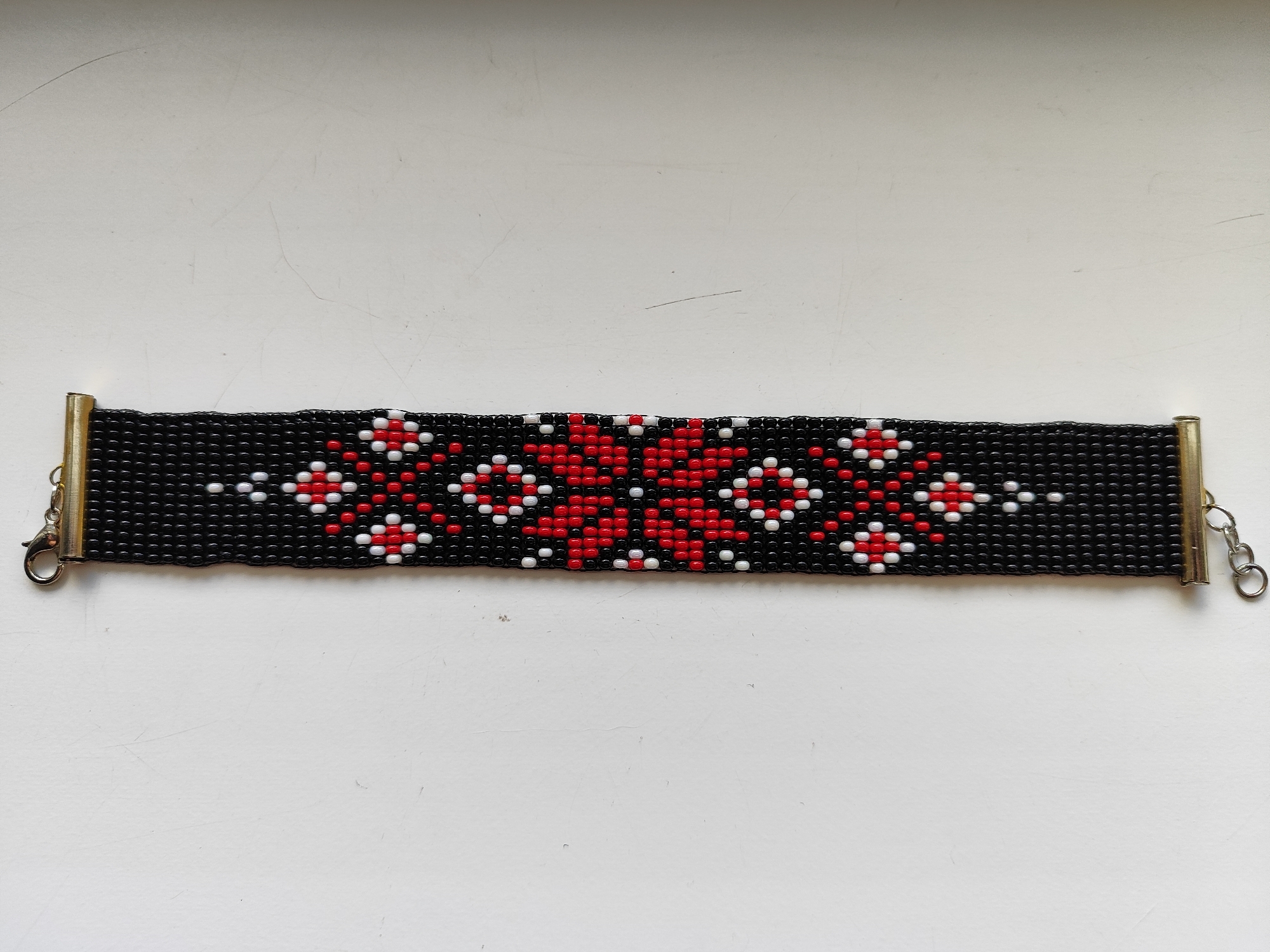 A beaded bracelet is lying on a flat, white surface. The bracelet is rectangular in shape with a black background and a repeating pattern of red and white floral designs. The ends of the bracelet are capped with gold-colored metal pieces and each end has a silver clasp attached. The location is Unknown, Unknown.