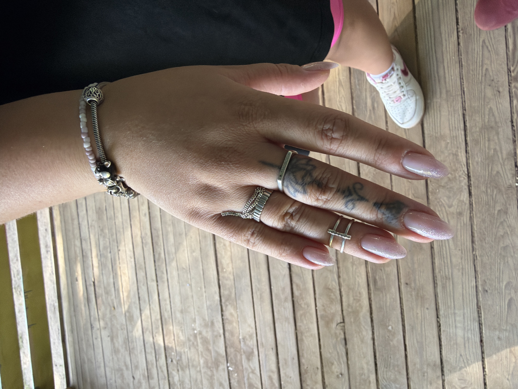 A person is displaying their hand, which has several rings on the fingers and a bracelet on the wrist. There are tattoos visible on the hand. In the background, a child's foot with a white sneaker is visible, along with wooden planks, possibly a porch or deck. The location is unspecified and there is no visual indication that the location is Chaikovsky, Russia.