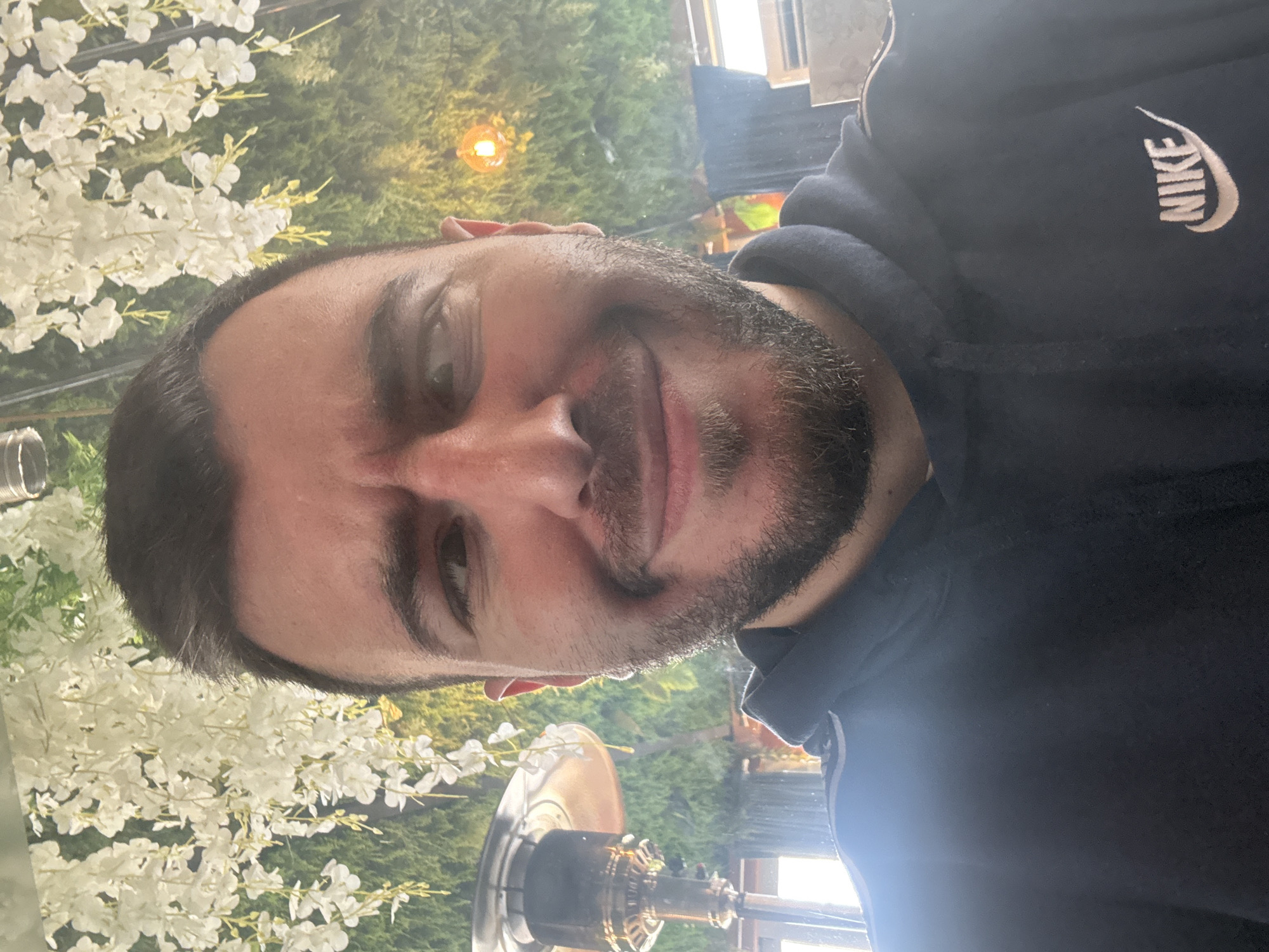 A man with short dark hair and a beard is posing for a selfie. He is wearing a dark blue hoodie with the Nike logo on the right shoulder. The background features white flowering branches and green foliage, with a warm-toned light fixture visible. In the foreground is a brass-colored outdoor heater. Based on the style of the setting, it's plausible the image was taken at an outdoor patio or restaurant in Sofia, Bulgaria, although specific details to confirm the exact location are absent.