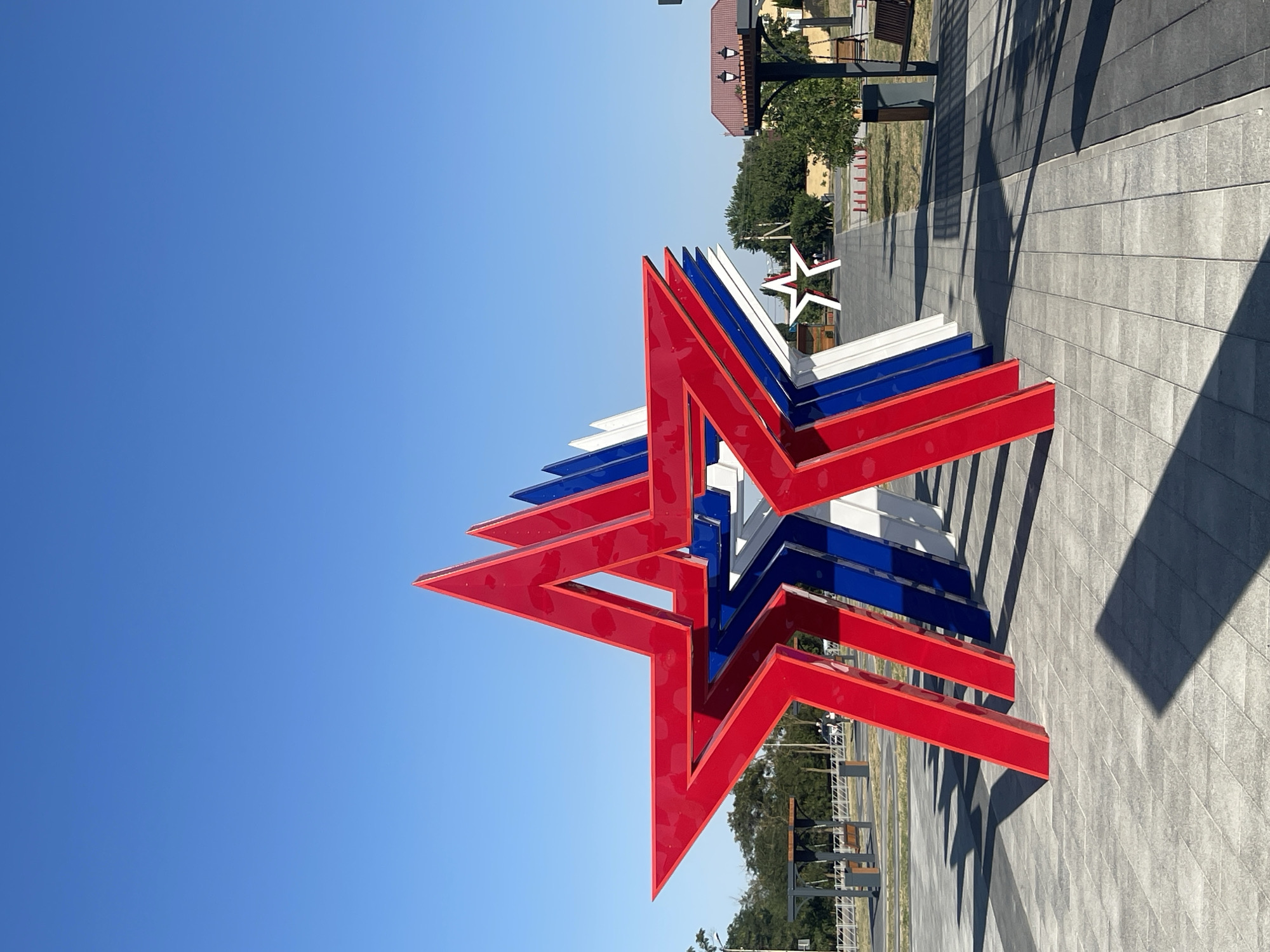A row of large, three-dimensional stars, painted in the colors red, white, and blue, are displayed on a paved plaza under a clear blue sky. The stars are positioned at an angle, creating a sense of depth and perspective. The foreground features the shadow of the closest red star cast across the grey tiled pavement. In the background, trees and buildings are visible, including a structure with a red roof and an ornate lantern. Benches and what appear to be picnic tables are also present, suggesting a public park or recreational area. The scene is located in the city of Budyonnovsk, Russia, likely at a memorial or public art installation. There are no people visible in the image.