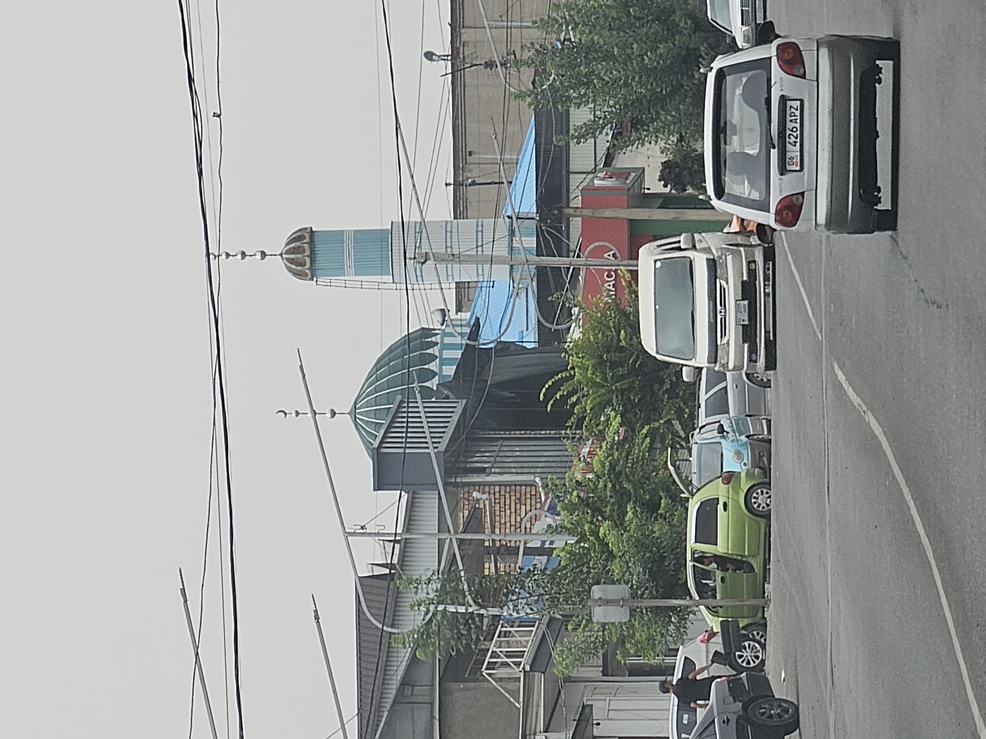 The image depicts a street scene in Özgön, Kyrgyzstan, featuring a prominent mosque with a green dome and a tall, light blue minaret. Several vehicles are visible on the asphalt road, including a silver sedan in the foreground with the license plate "G6 426 APZ", a white van, and a lime green compact car with its doors open. A person is standing near the lime green car. The environment is urban, with buildings and trees lining the street, and overhead electrical wires are prevalent. The sky is overcast.