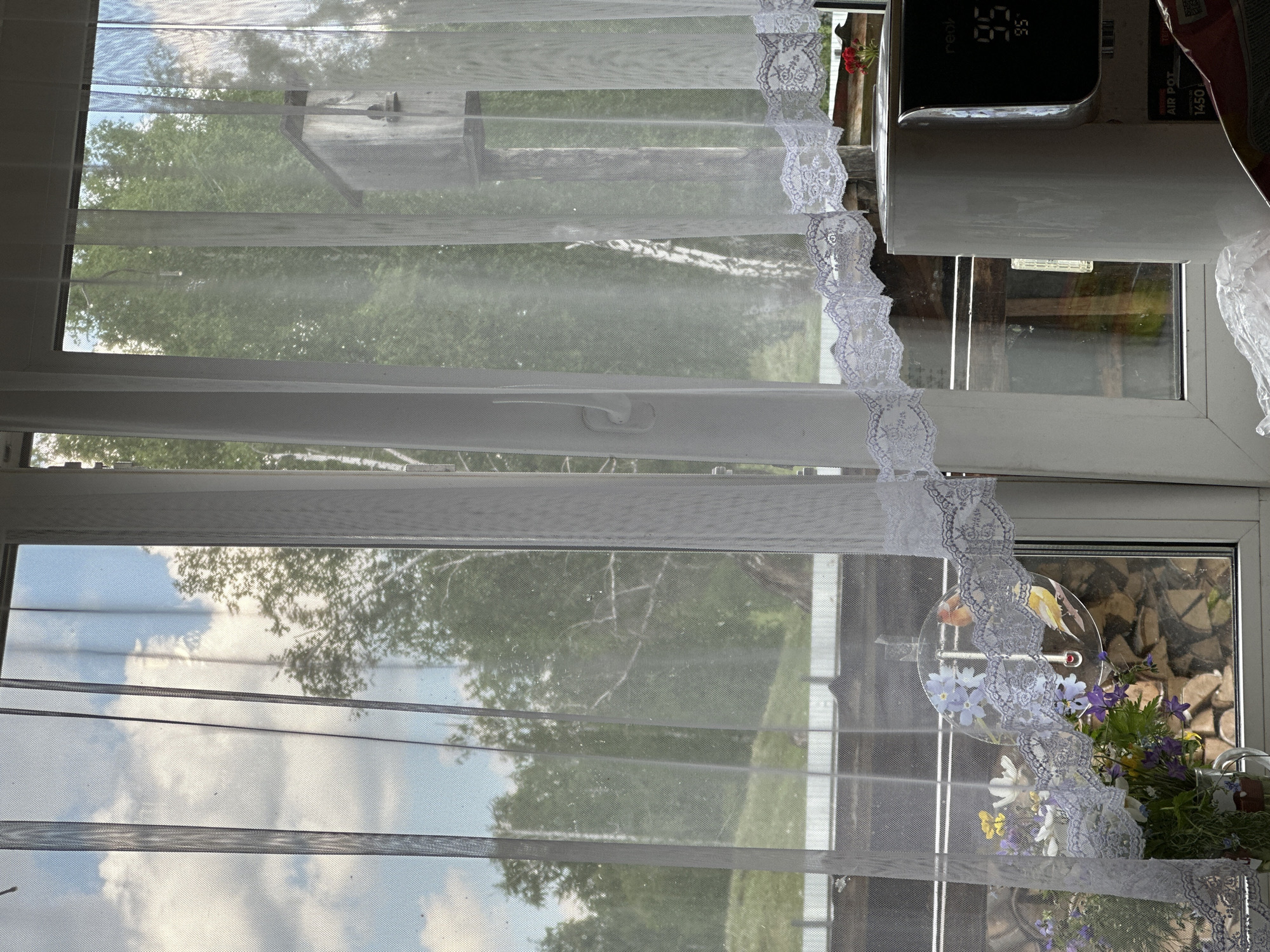 A daytime interior shot shows a window covered with a sheer white curtain with lace trim. Outside, trees and vegetation are visible, along with glimpses of the sky. A white air purifier and an air pot are partially visible on the right side of the window frame. A glass disc with painted flowers hangs in the window, partially obscured by the lace curtain. Below the disc, a bouquet of flowers in a glass vase is also visible. The image appears to be taken in an urban setting, as there are signs of buildings and structures outside the window. The location is Unknown, Unknown.