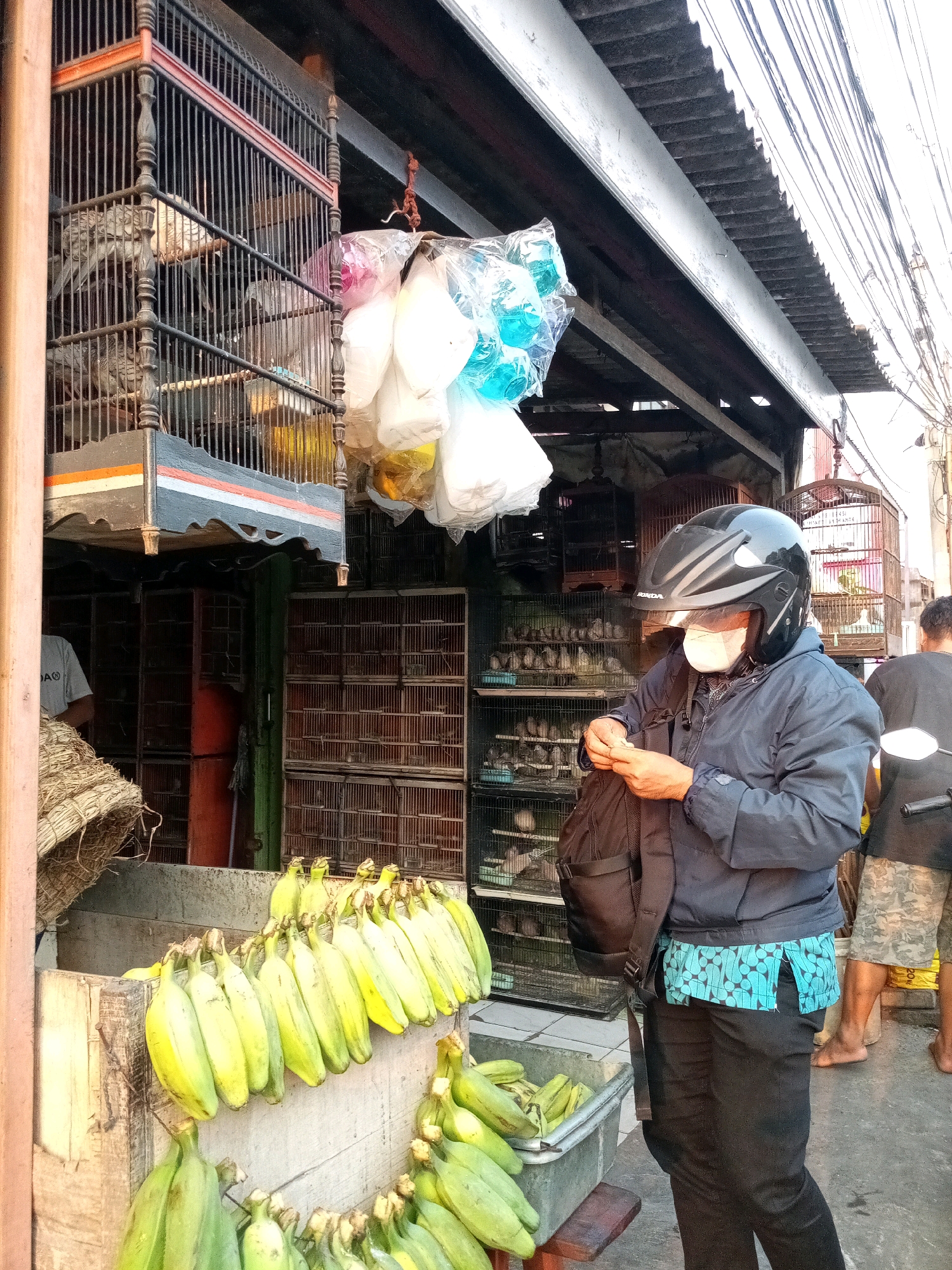 The image captures a street scene in Karangsatria, Indonesia, likely a market area. In the foreground, a person wearing a black helmet and a light blue patterned shirt with black pants stands facing right, handling what appears to be money. They have a black backpack slung over their left shoulder and a white mask covering their nose and mouth. To the left of this person is a display of green bananas arranged in bunches. Behind the bananas and the person, a shop is visible, with multiple cages stacked vertically containing small birds. A large, ornate birdcage hangs from the awning above the shop. In the background, another person with camouflage shorts and a black t-shirt is partially visible, facing away from the camera. The environment suggests an outdoor market with a simple, weathered awning providing shelter for the shop. The lighting indicates it is likely daytime.