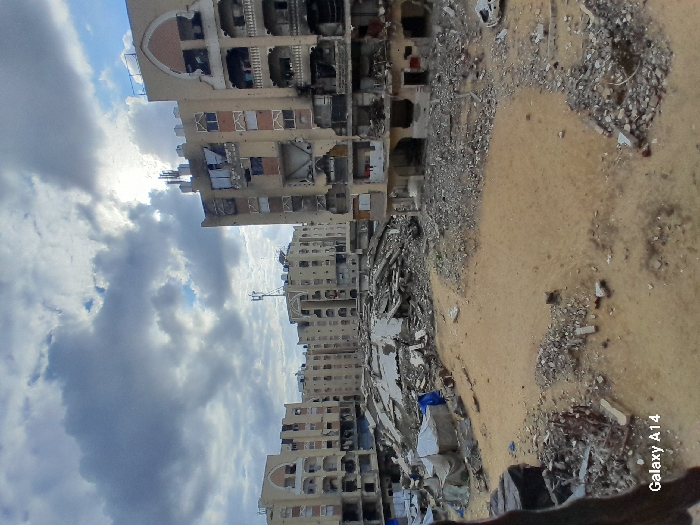 The image depicts an urban landscape in Khan Yunis, Palestinian Territory, characterized by widespread structural destruction under an overcast sky. Numerous multi-story residential-style buildings, typically five to eight stories tall, line the perimeter of the scene, all exhibiting severe damage. Many structures feature significant facade collapses, exposing interiors, shattered windows, and large holes. Burn marks and discoloration are visible on multiple building surfaces, particularly on the prominent building to the right, which shows extensive damage across its lower and middle floors, with exposed concrete and twisted rebar. Architectural details such as arched balcony openings and decorative elements are discernable on some less obliterated sections.
The central ground area consists of exposed, light brown earth and sand, heavily strewn with concrete rubble, fragmented building materials, and debris. Large, irregular mounds of pulverized concrete and other structural remnants are prominent in the middle ground, particularly towards the left. A blue tarpaulin and what appears to be a makeshift tent or shelter are partially visible amidst the debris piles on the left. On the far right, a damaged white vehicle, possibly a car chassis, is partially buried in rubble. The sky above is largely covered by dense, grey clouds, with patches of brighter sky suggesting an overcast or partially cloudy day. No individuals or specific actions are discernible within the frame. A "Galaxy A14" watermark is present in the lower-left corner.