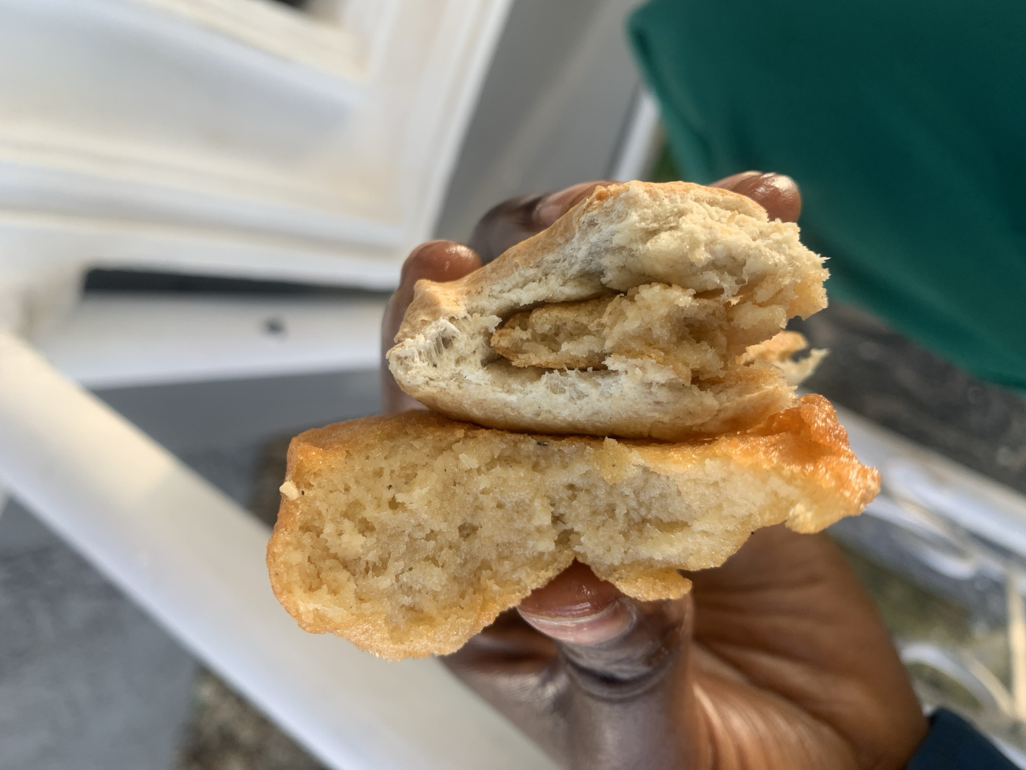 A close-up shot shows a pair of hands holding a fried dough pastry that has been broken in half. The pastry has a golden-brown exterior and a soft, airy interior with visible pockets. The hands appear to be a person of color, with manicured fingernails. The background is blurred, with hints of white and gray structures, suggesting an outdoor or well-lit indoor setting. There is no identifiable information about the location in Gura Top, Nigeria.