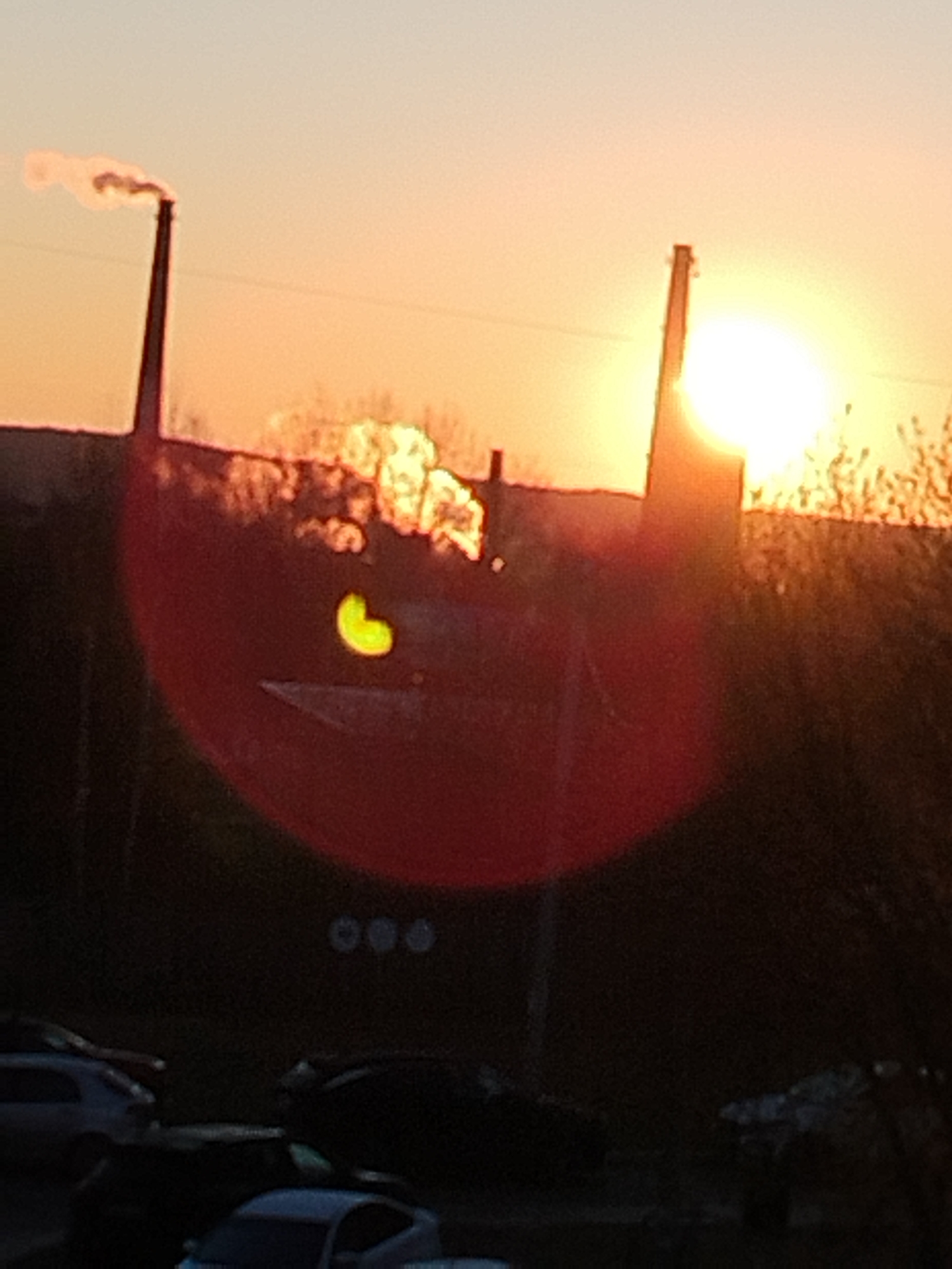 The image shows a sunset scene in Chaikovsky, Russia. Two industrial chimneys dominate the skyline. The left chimney is emitting smoke. A bright, round sun is visible behind the right chimney. The foreground is dark and shows silhouettes of cars parked in a lot. The image has a strong red lens flare. There is also a yellow heart or kidney-bean shaped object in the red lens flare. The location is likely an area with industrial infrastructure near a residential or commercial zone in Chaikovsky, Russia.