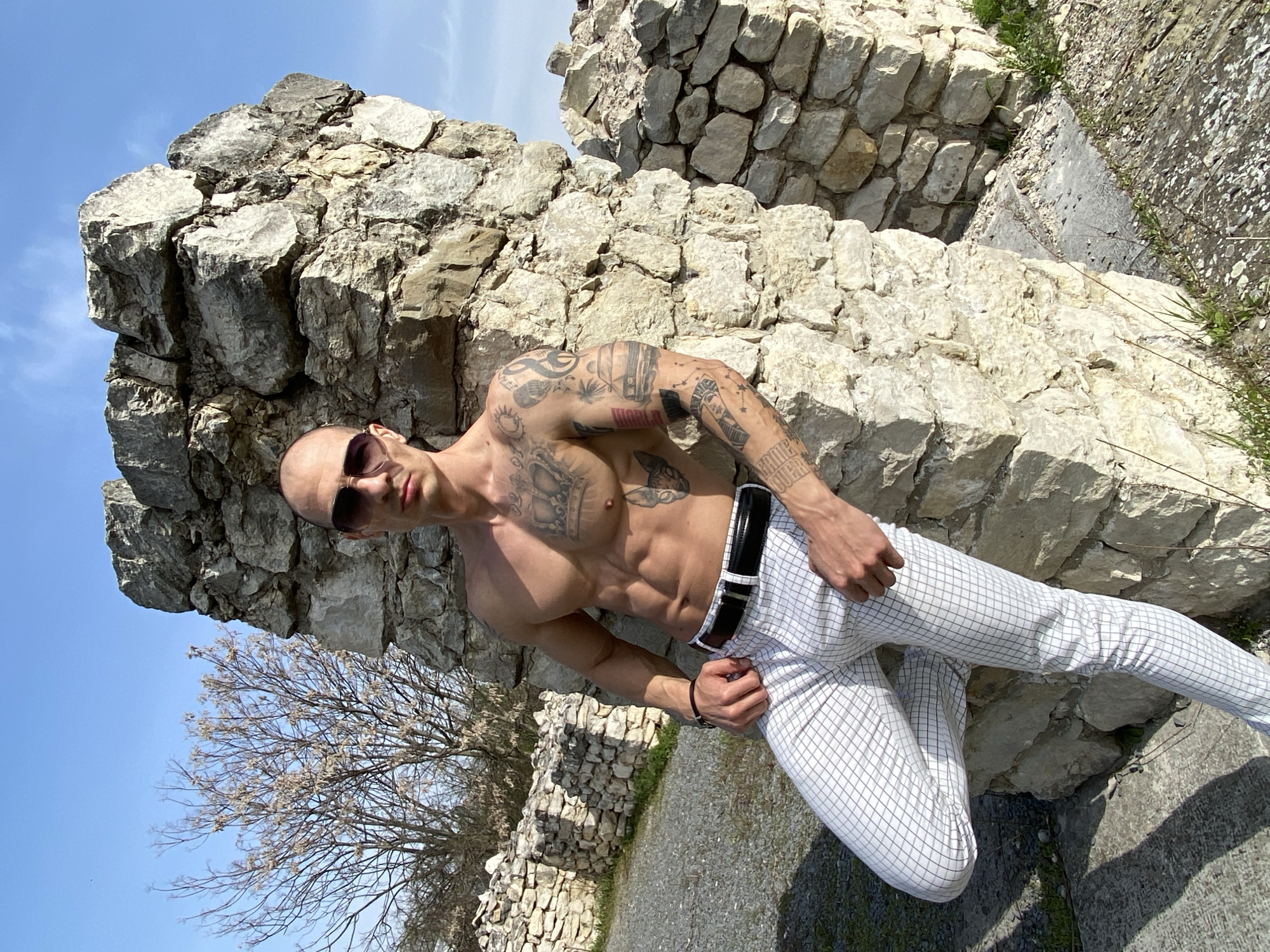 A bald, muscular man with sunglasses and multiple tattoos is posing outdoors, leaning against a light-colored stone structure. He is bare-chested and wearing checkered white pants with a black belt. The background includes a blue sky, a bare tree, and other stone structures. The location is the Kaylaka Park in Pleven, Bulgaria.