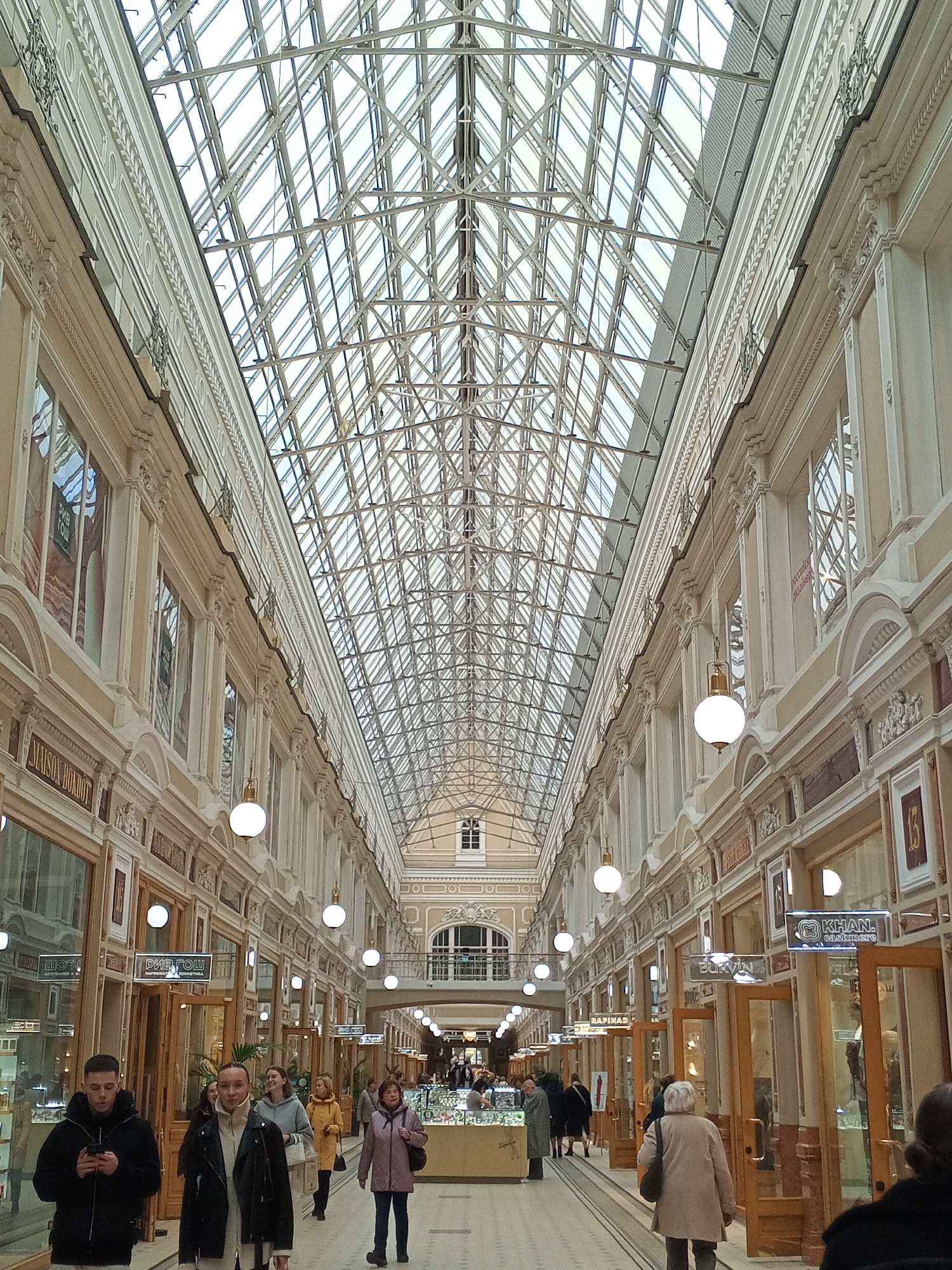 The image shows an interior view of the Passage in Saint Petersburg, Russia. The long, narrow arcade is lined with shops on both sides. The ceiling is a complex lattice of metal beams and glass panels, allowing natural light to flood the space. White globe lights hang from the ceiling at regular intervals, illuminating the shops and the walkway. People are walking through the passage, some looking at their phones, some window shopping, and others simply passing through. Most are wearing casual clothing appropriate for a city environment. There are shop signs visible above some of the storefronts, indicating various types of businesses. The architecture is ornate, with decorative moldings and embellishments along the upper walls and around the windows. The overall impression is of a bustling, elegant shopping arcade with a distinctive architectural style.