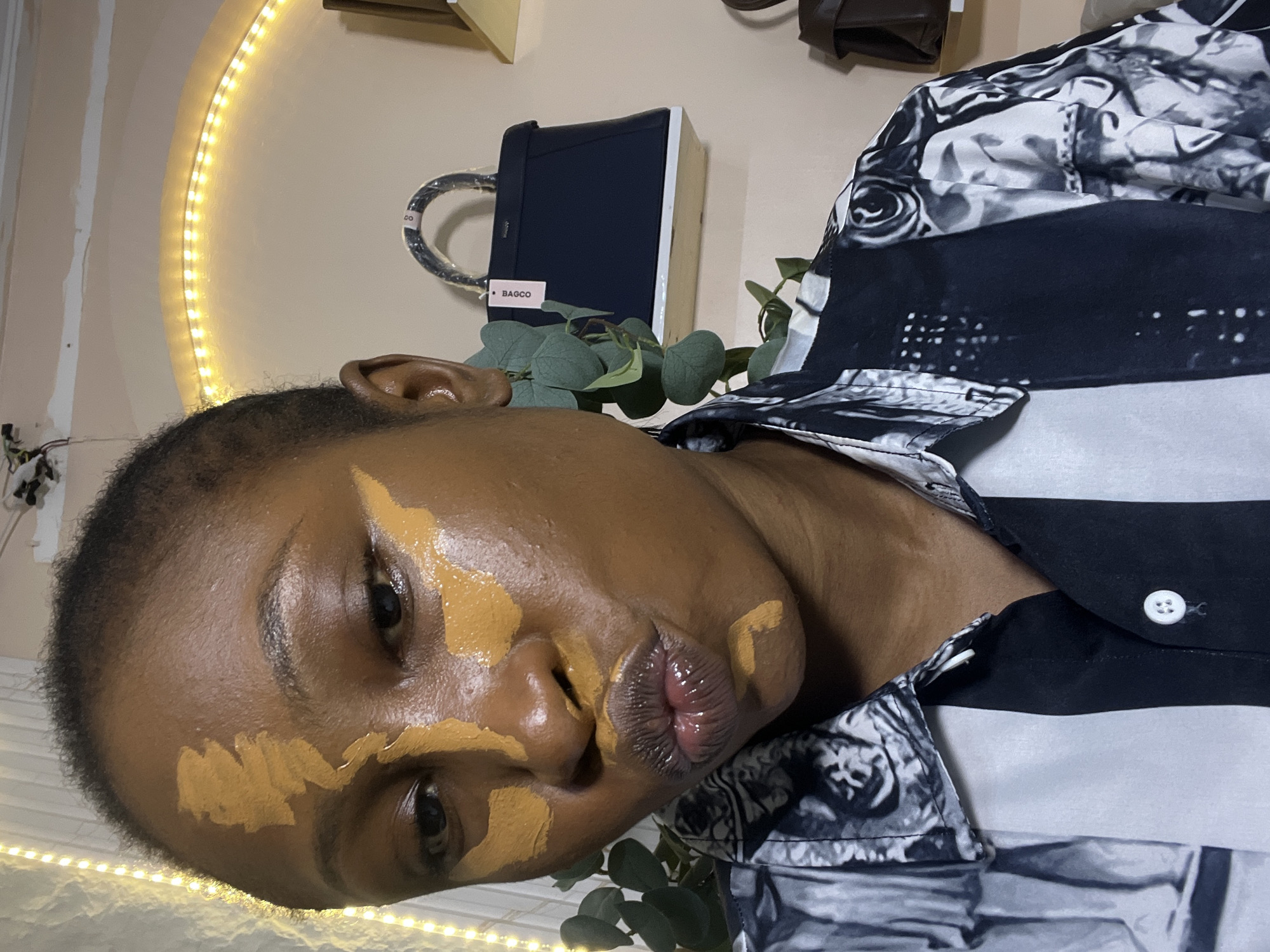 This is a vertical, close-up photograph of a Black person applying foundation to their face. The person is looking towards the camera, with foundation applied in patches across their cheeks, forehead, and nose. Their lips are slightly parted, revealing a glossy texture. The person is wearing a black and white patterned button-up shirt.
In the background, a circular array of warm-toned LED lights is visible on the wall, suggesting an indoor setting, possibly a room. To the right of the person's head, a dark blue, rectangular object with a silver handle is partially visible, with a pink label that reads "BAGCO". Some green foliage is also present in the background. The lighting appears to be artificial, with a warm glow cast by the LED lights. The overall impression is that of a self-application of makeup, captured in a casual, personal moment within a home environment in Abuja, Nigeria.