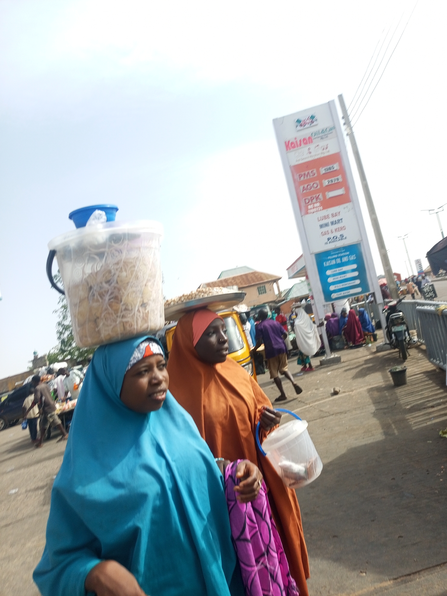The image shows two women in hijabs walking in Gombe, Nigeria. The woman on the left wears a blue hijab and carries a clear plastic container of food on her head. The woman on the right wears an orange hijab, holds a clear plastic container in her hand, and has a purple and pink patterned cloth draped over her arm. In the background, there is a "Kaisan Oil and Gas" sign. Other people and vehicles are visible in the background.