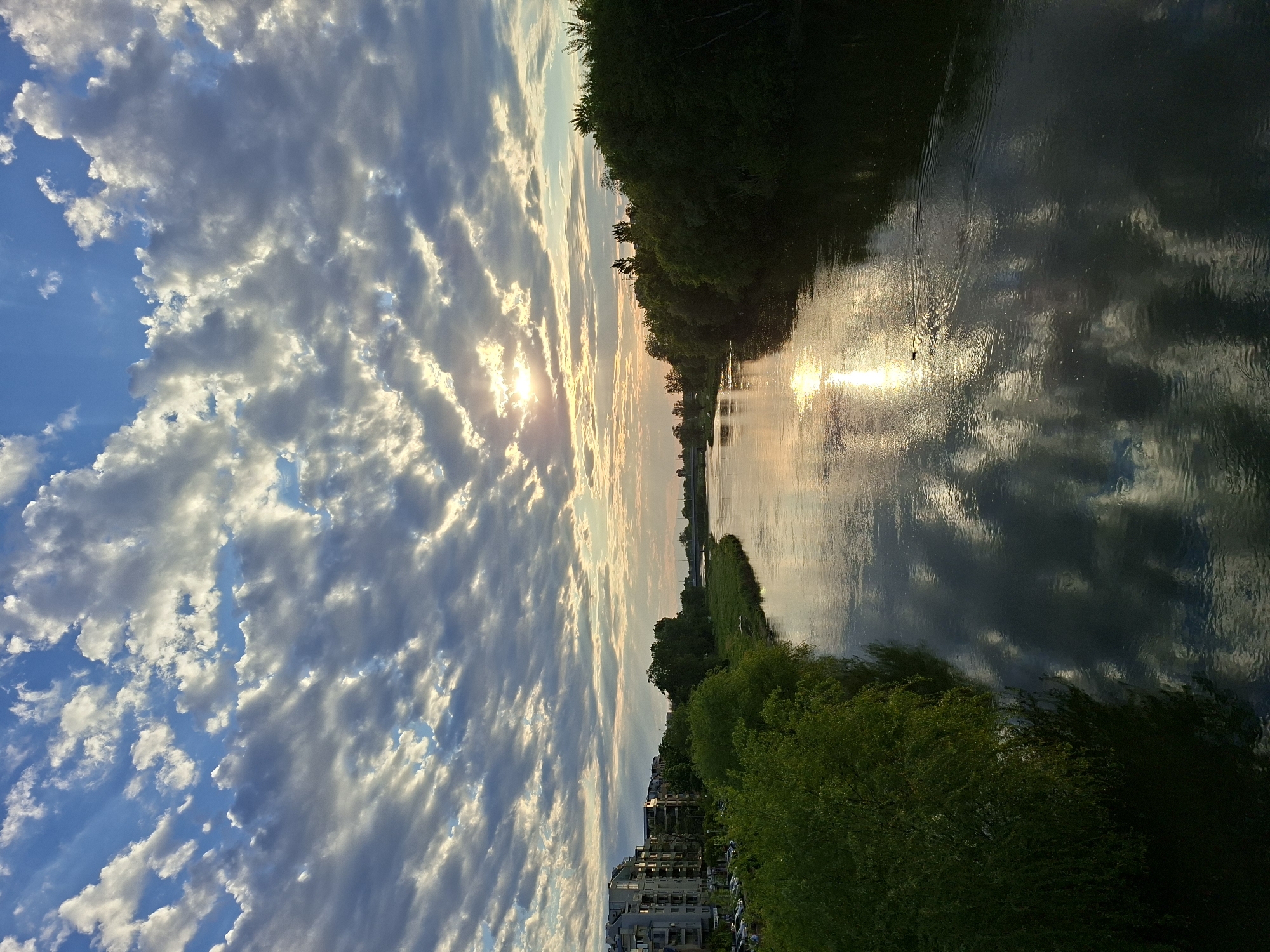 The image presents a view of the Maritsa River in Plovdiv, Bulgaria, at sunset. The river reflects the sunlight and the sky. On the banks are trees and vegetation. Buildings are also visible on one side of the riverbank. The sky is filled with clouds.