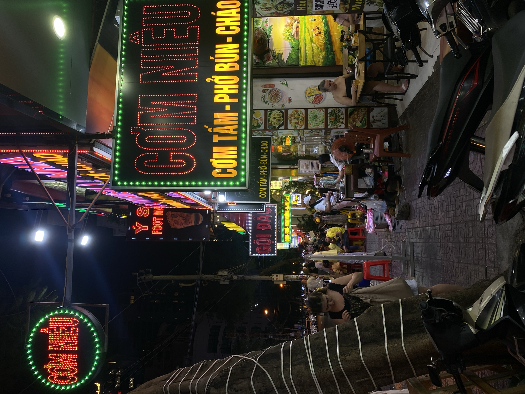 This is a nighttime street scene in Nha Trang Ward, Vietnam, characterized by vibrant neon signs advertising food establishments. The primary focus is on a large, vertical neon sign that reads "COM NIEU" in red lettering, with "CƠM TẤM - PHỞ BÚN - CHÁO" in yellow below it. Another similar sign, "COM NIÊU," is visible with a green circular border.
The scene depicts a bustling street-level restaurant or food stall area. People are seated at tables, some appearing to be dining or socializing, while others are engaged in activities related to the establishment, such as serving or preparing food. Motorbikes are parked along the sidewalk, a common sight in Vietnamese street scenes. The lighting is predominantly from the neon signs and streetlights, casting a warm, inviting glow on the surroundings.
Other visible text includes "YS FOOT MA," potentially a sign for a foot massage service, and other fragmented Vietnamese text, such as "GỌI ĐAU," "Hàu," and "Mực," suggesting a variety of food options. The overall impression is one of a lively, urban environment where food and commerce are central. The time of day is clearly night, with the darkness of the sky contrasting sharply with the illuminated signs. The weather appears to be clear, with no indication of rain. The presence of numerous people and the active dining setting suggest a popular local spot.